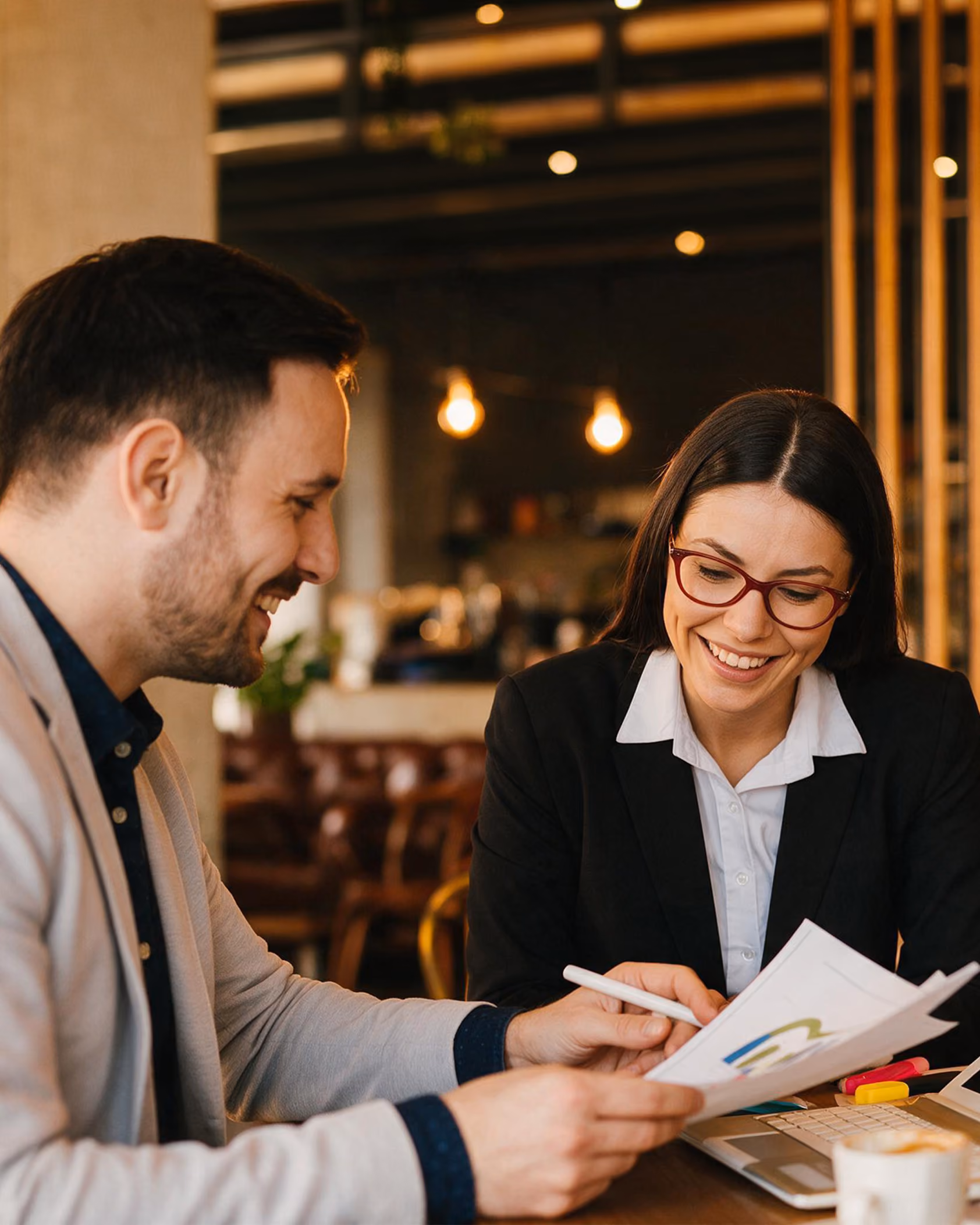 Smiling man and woman in business attire reviewing a document with charts together in a modern, warmly lit office setting.