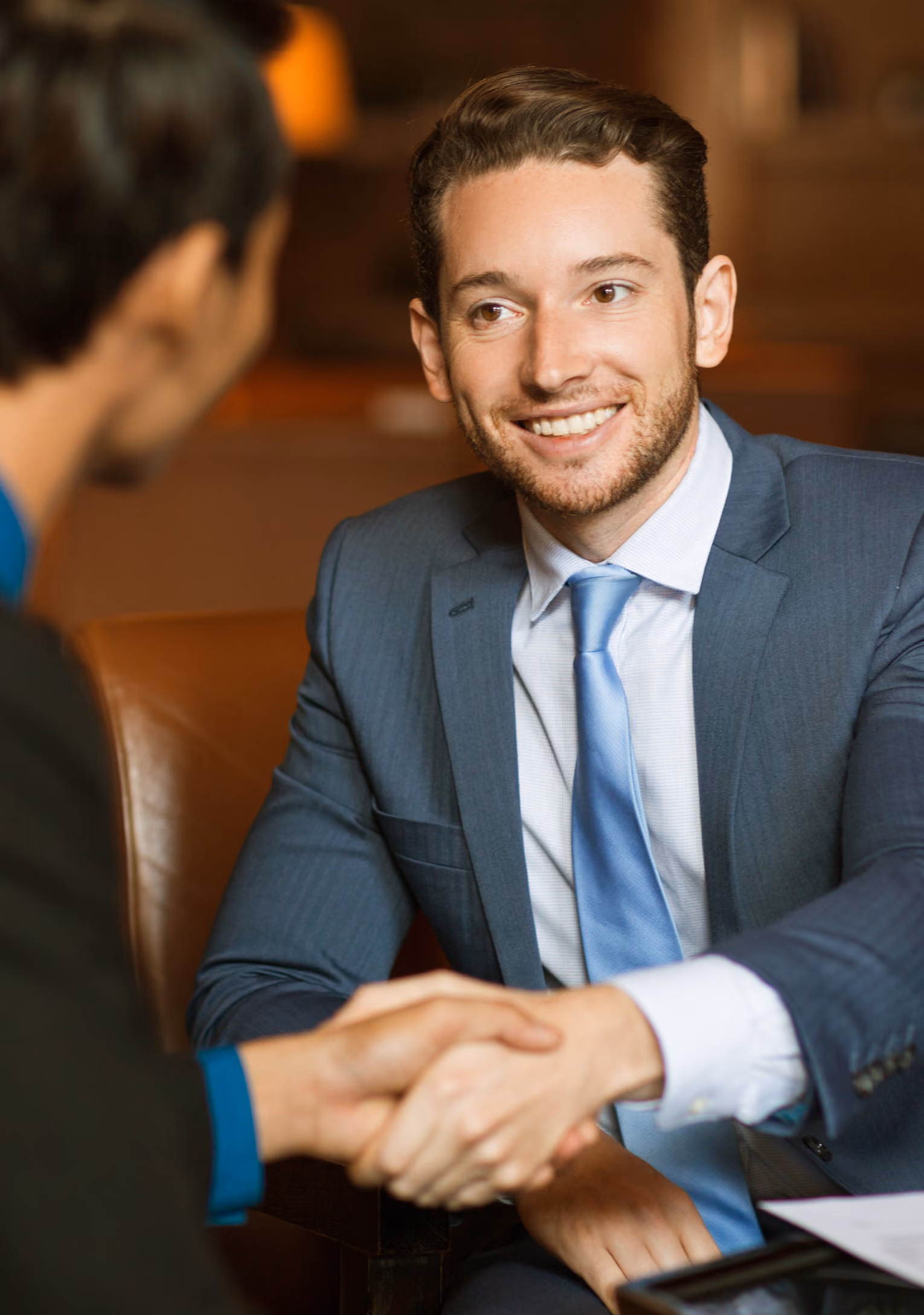 A smiling man in a blue suit and light blue tie shaking hands with another person during a meeting.