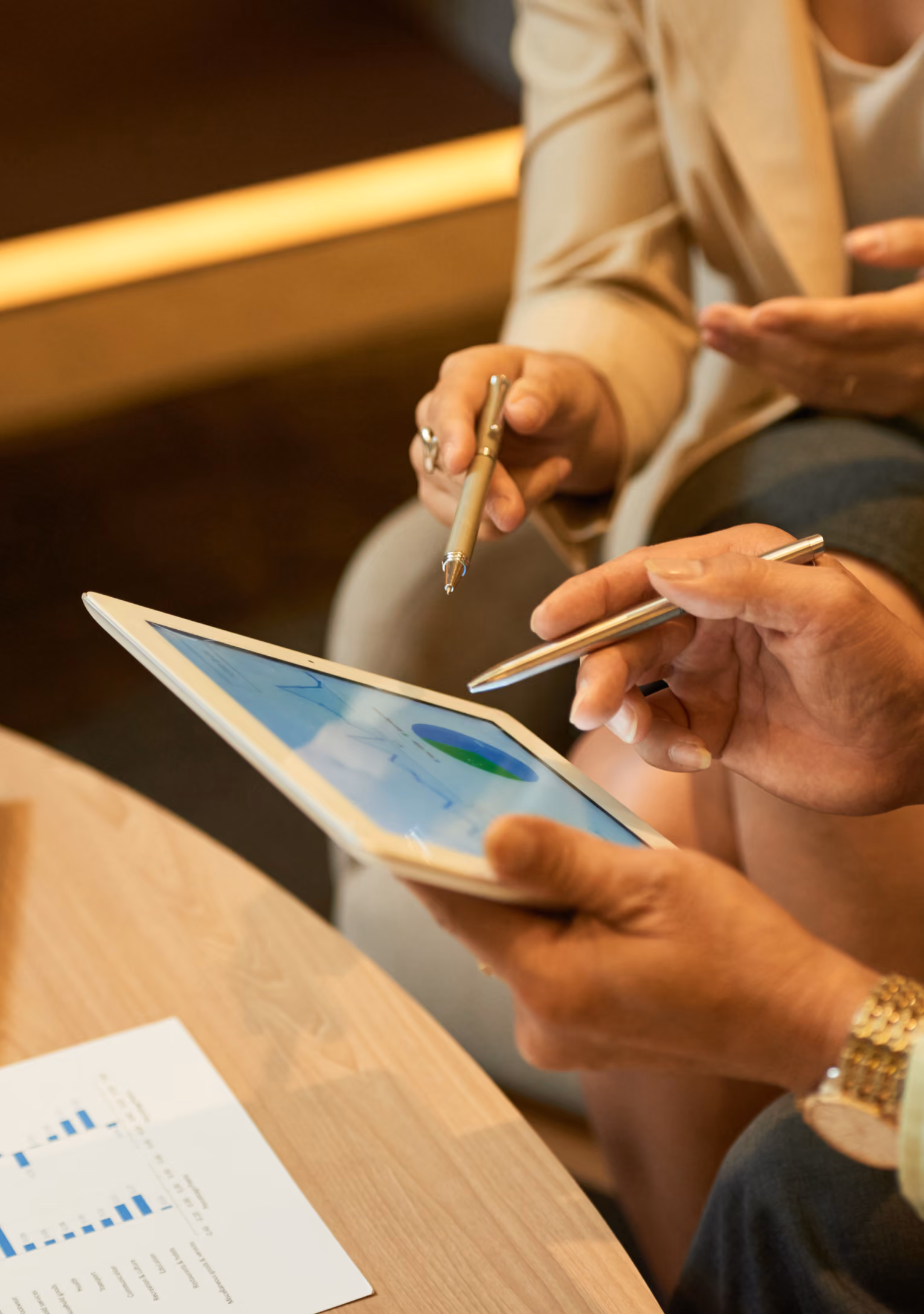 Two people sitting and discussing data shown on a tablet screen, one pointing at the screen with a pen, with a printed bar chart on the table nearby.