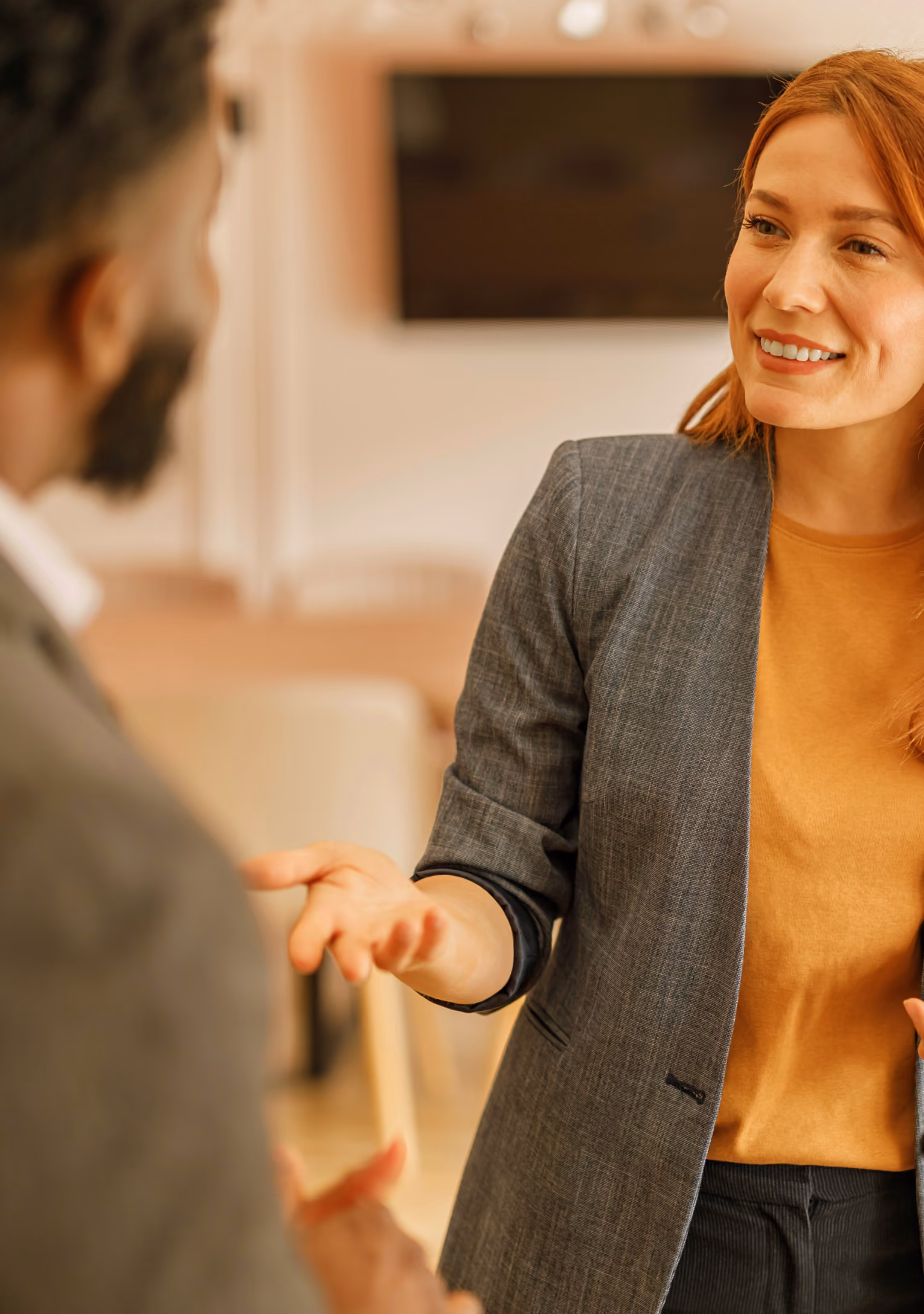 A red-haired woman in a gray blazer and mustard shirt engaging in a friendly conversation with a man who is partially visible and blurred in the foreground.