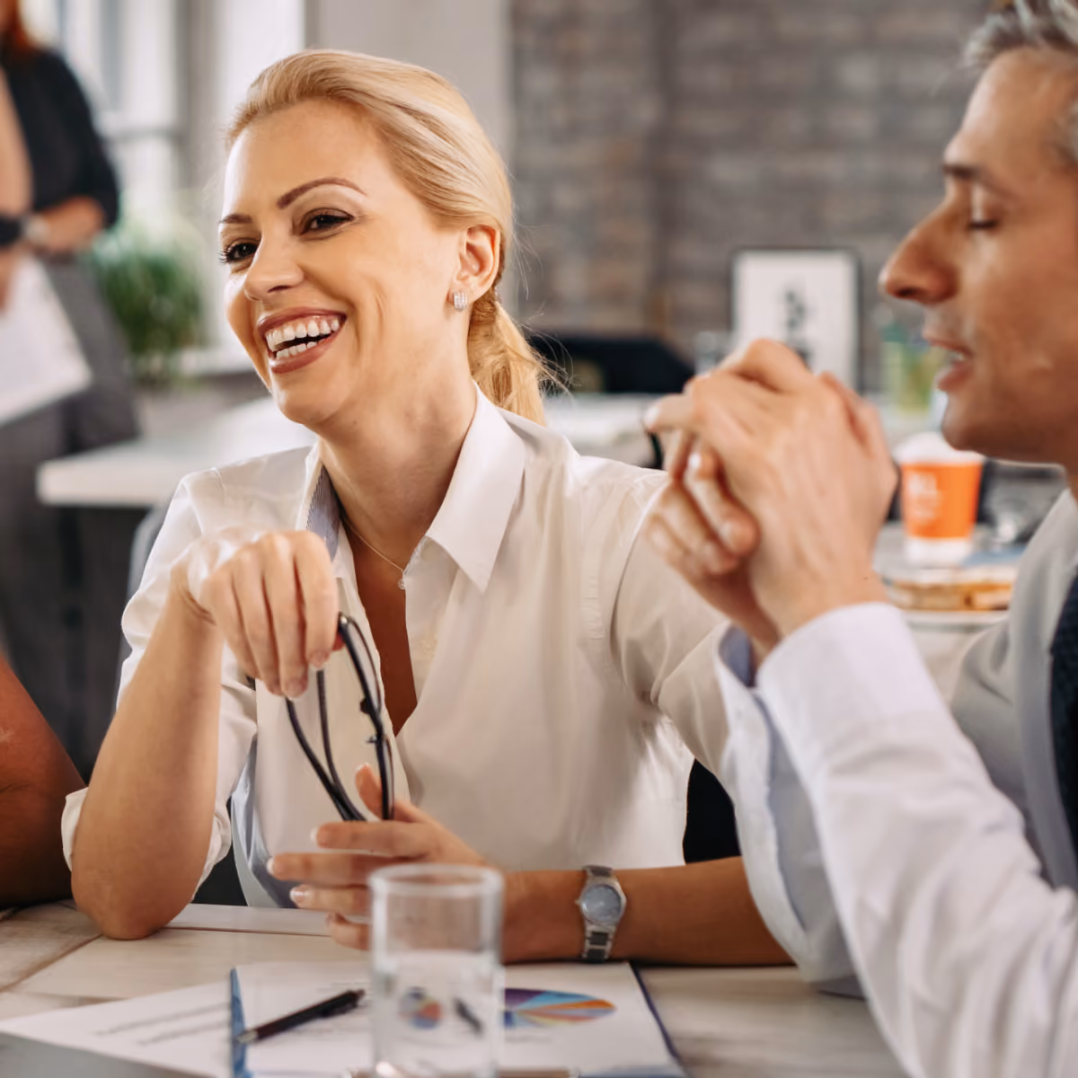 Smiling woman holding eyeglasses and sitting at a table with documents and a glass of water, engaged in conversation with a man beside her.