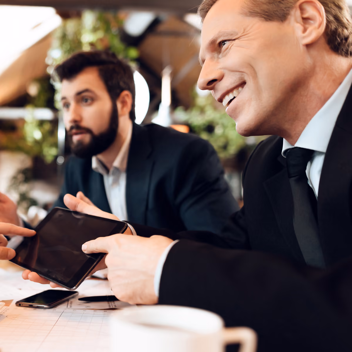 Two men in business attire sitting at a table; one man is smiling and holding a tablet while the other looks on attentively, with a coffee cup and smartphone on the table.