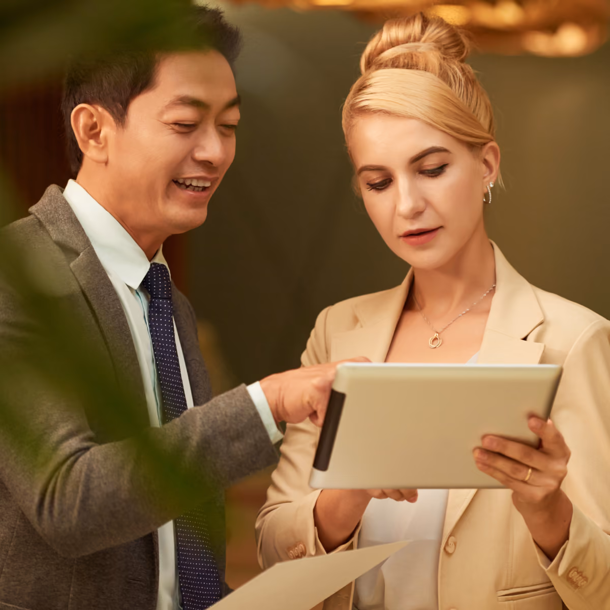 Two business professionals, a man in a suit and tie and a woman in a beige blazer, looking at a tablet together, with the man pointing at the screen and the woman holding a document.