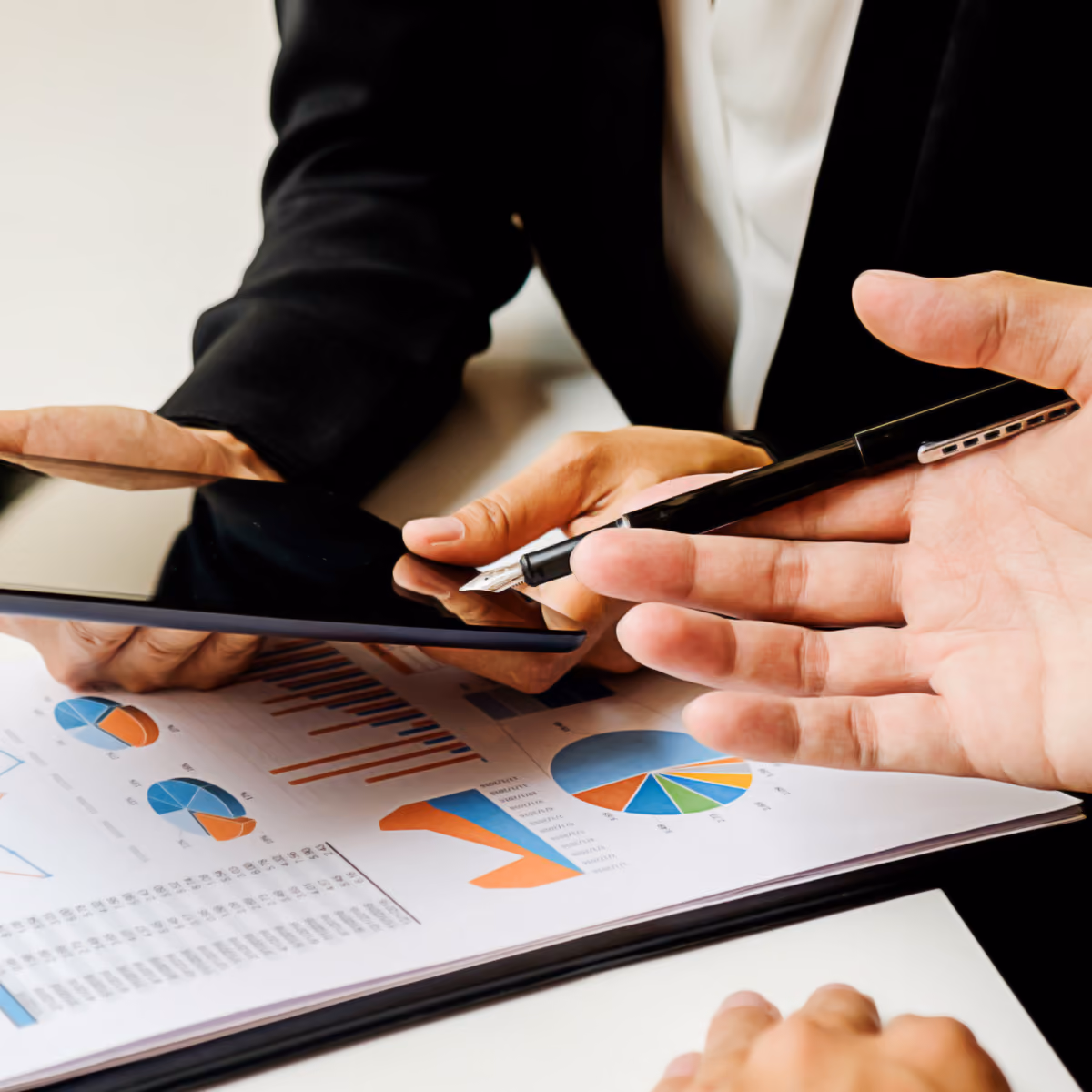 Two people in business attire analyze financial charts showing pie and bar graphs while using a tablet and a pen during a meeting.