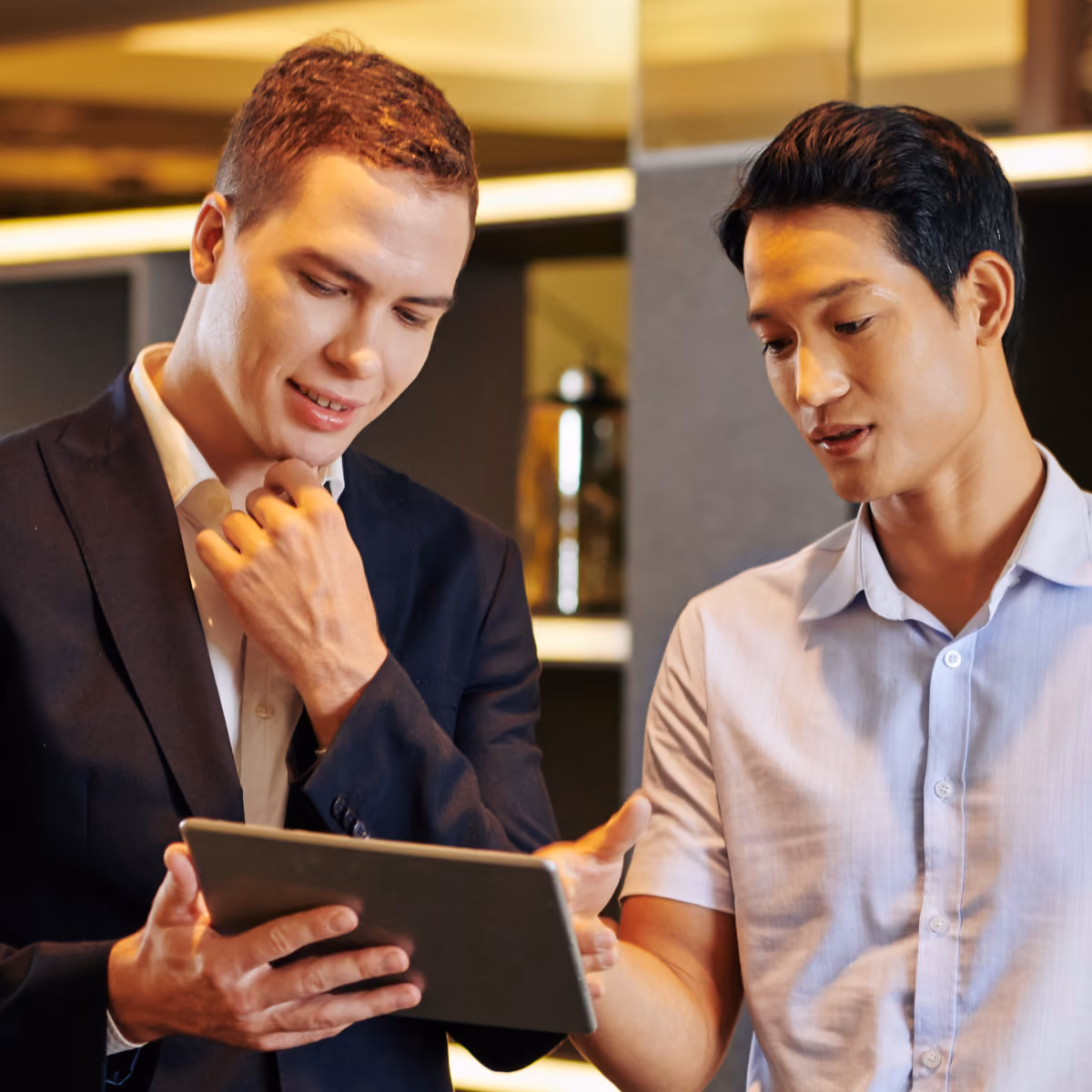 Two men in business attire looking at and discussing content on a tablet in a modern office setting.