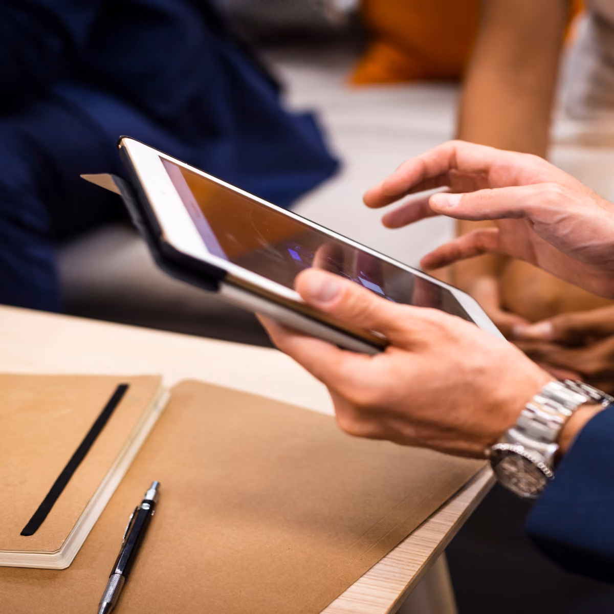 Person holding and interacting with a tablet device over a wooden table with a notebook and pen nearby.