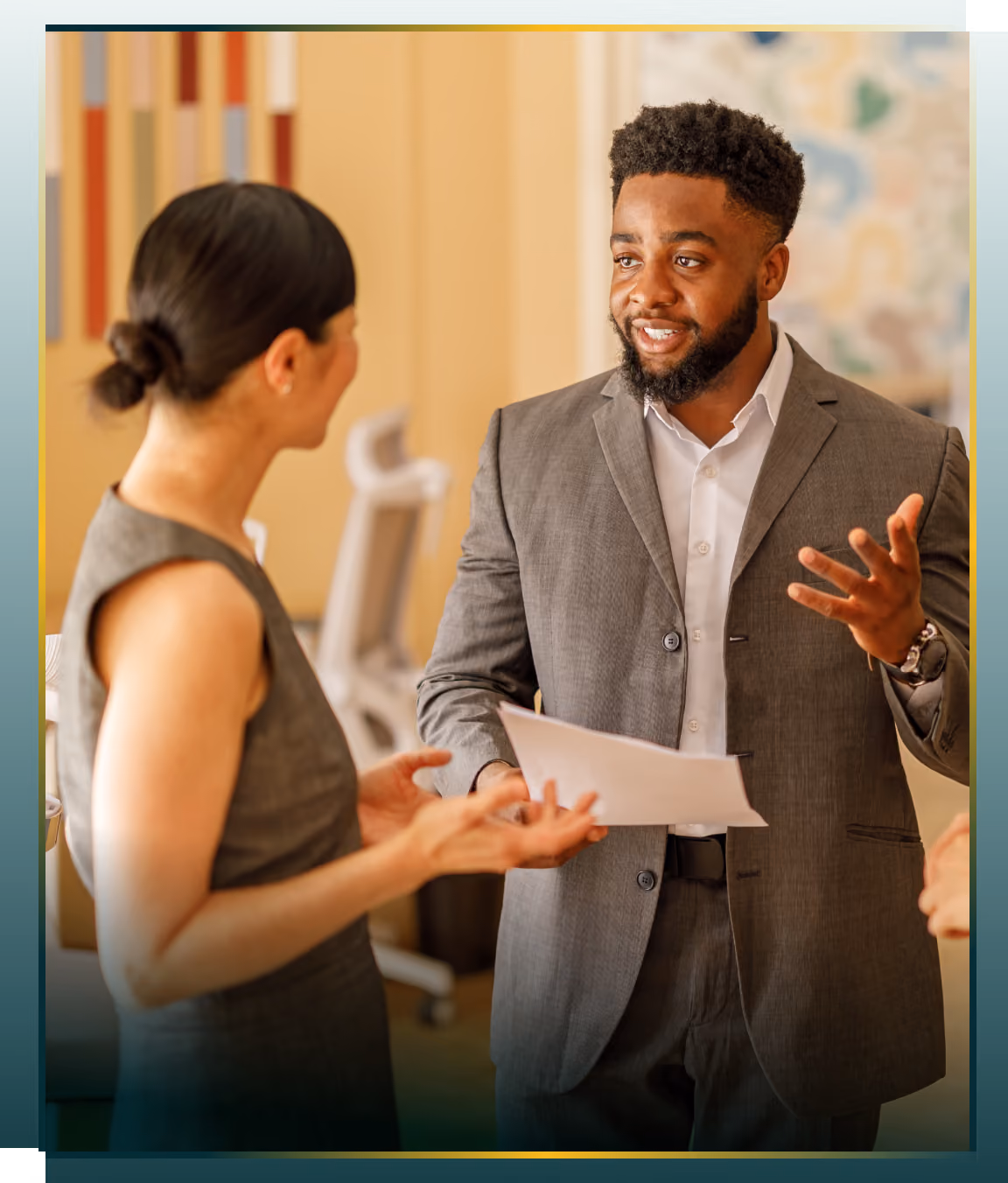 A man in a gray suit holding papers is talking and gesturing with his hand to a woman in a sleeveless dress in an office setting.