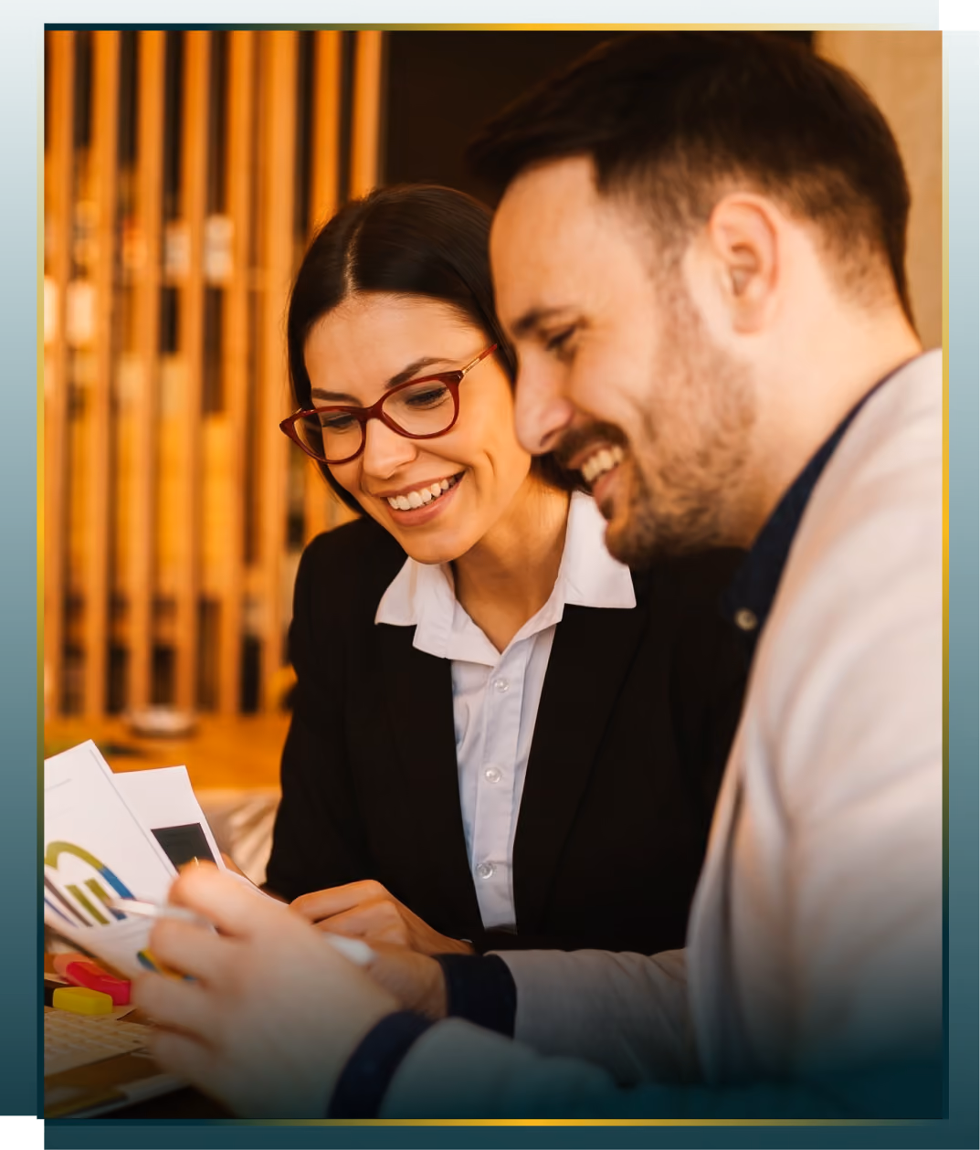 Two business professionals smiling and looking at documents together in an office setting, with a wooden slat background.