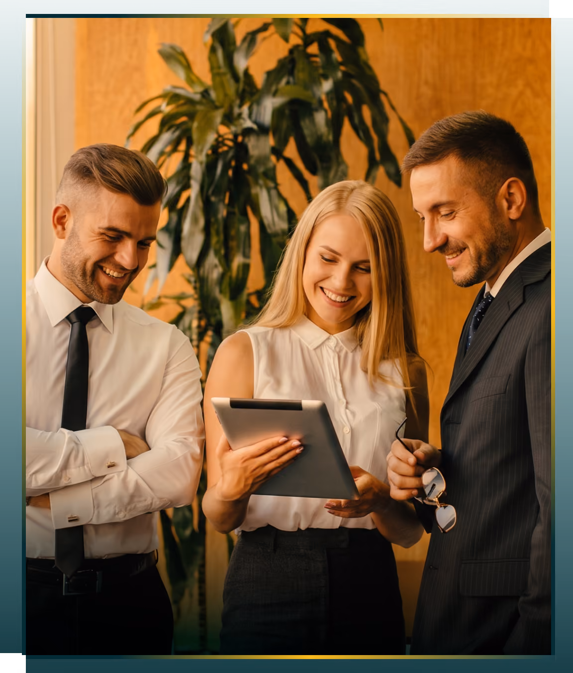 Three business professionals, two men and one woman, smiling and looking at a tablet device together in an office setting with a large plant and wood panel background.