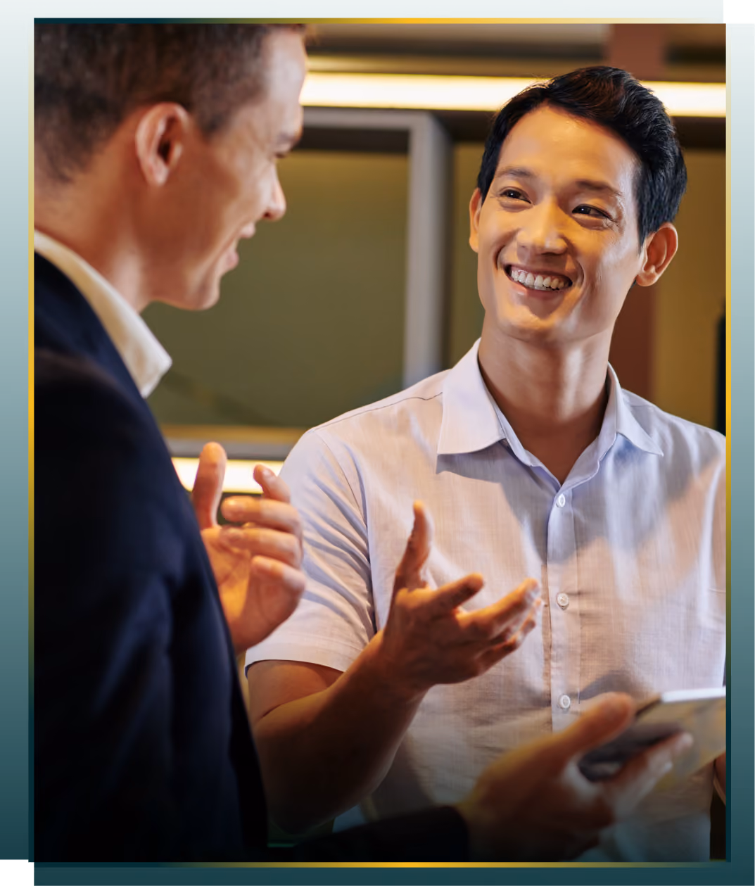Two men engaged in a friendly discussion, one smiling warmly while gesturing with his hands, the other holding a tablet and listening attentively.