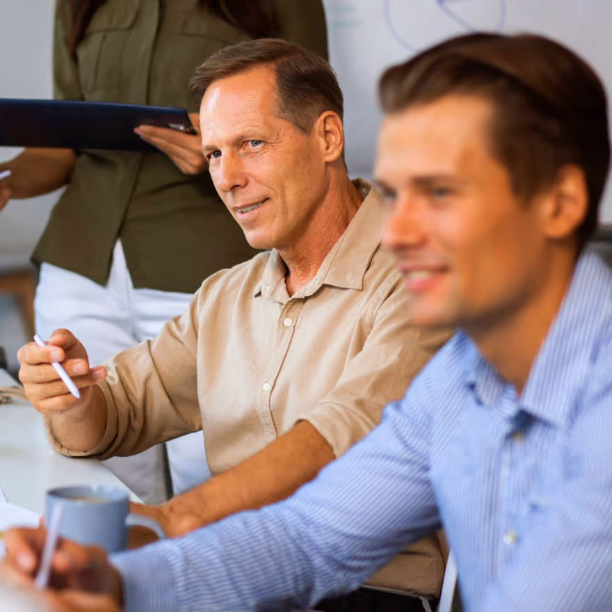 Two men sitting at a table during a meeting; one man in a beige shirt looks attentively while holding a pen, and the other man in a blue striped shirt is blurred in the foreground with a coffee mug nearby.
