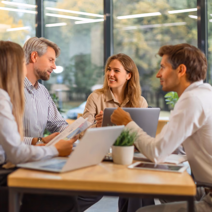 Four colleagues sitting around a table in an office with large windows, engaging in a discussion with laptops and documents.