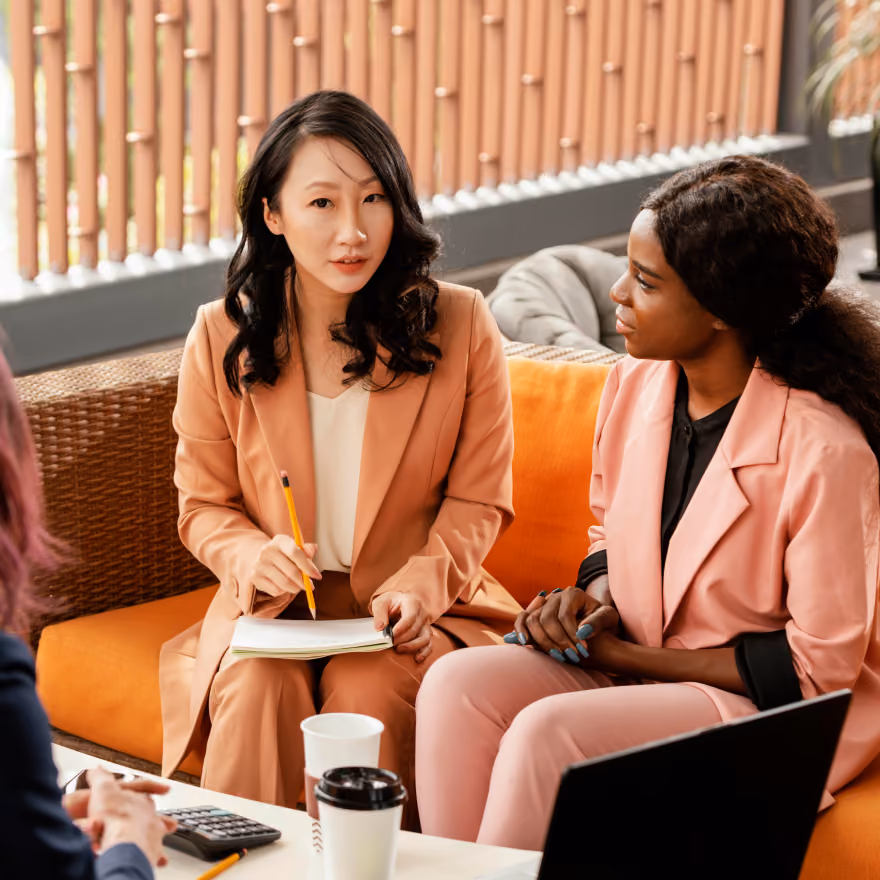 Two women in professional attire, one holding a pencil and notebook, engaged in conversation while sitting on an orange couch in a modern office setting.