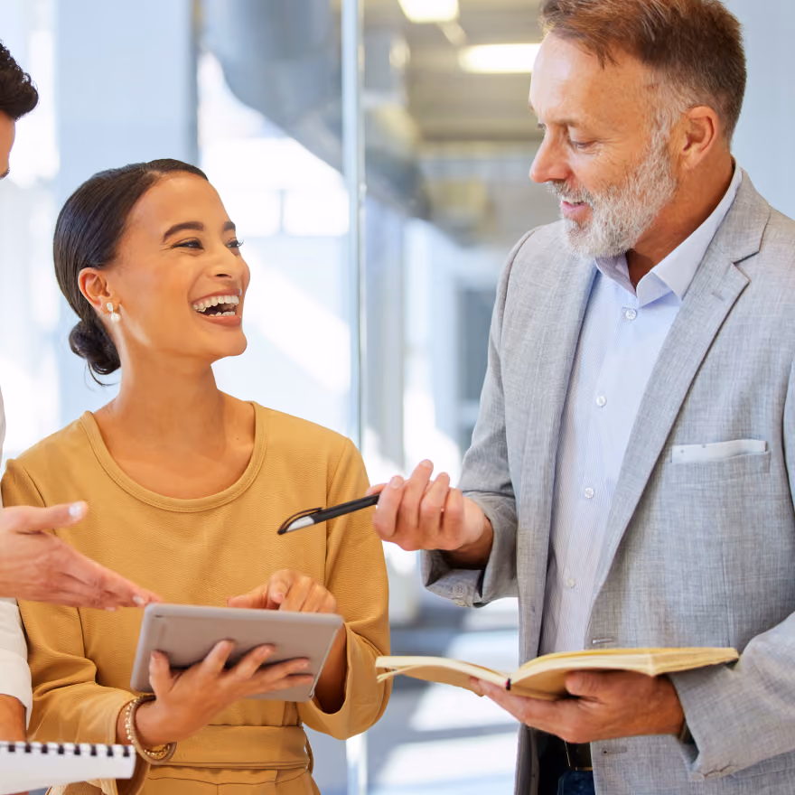 Three colleagues in a bright office engaged in a discussion; a smiling woman in a yellow top holds a tablet, a man in a gray suit holds a book and pen, and a third person gestures with one hand.