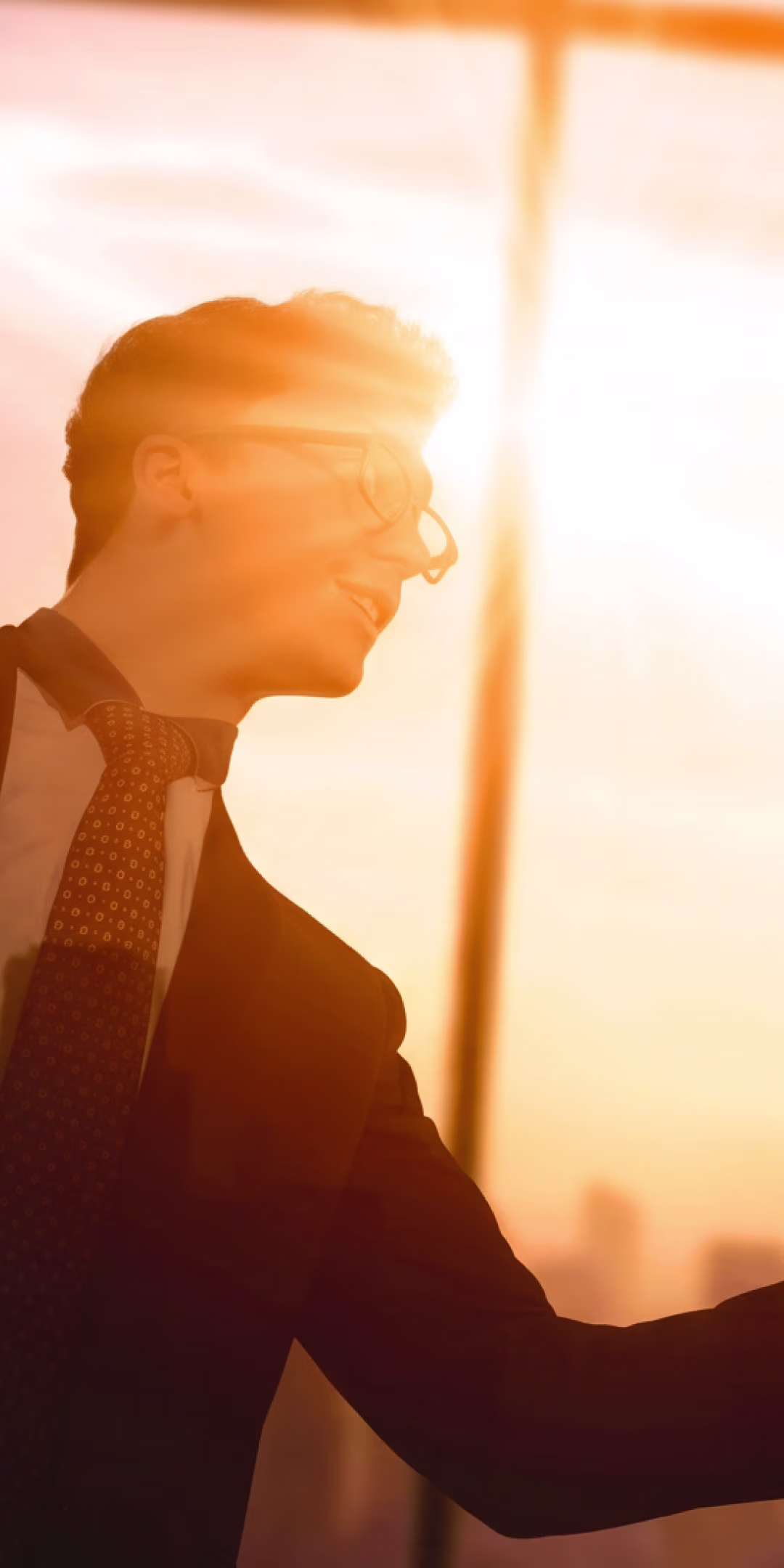 Man wearing glasses and a suit with a patterned tie, smiling and shaking hands, backlit by a bright golden sunset through a large window.