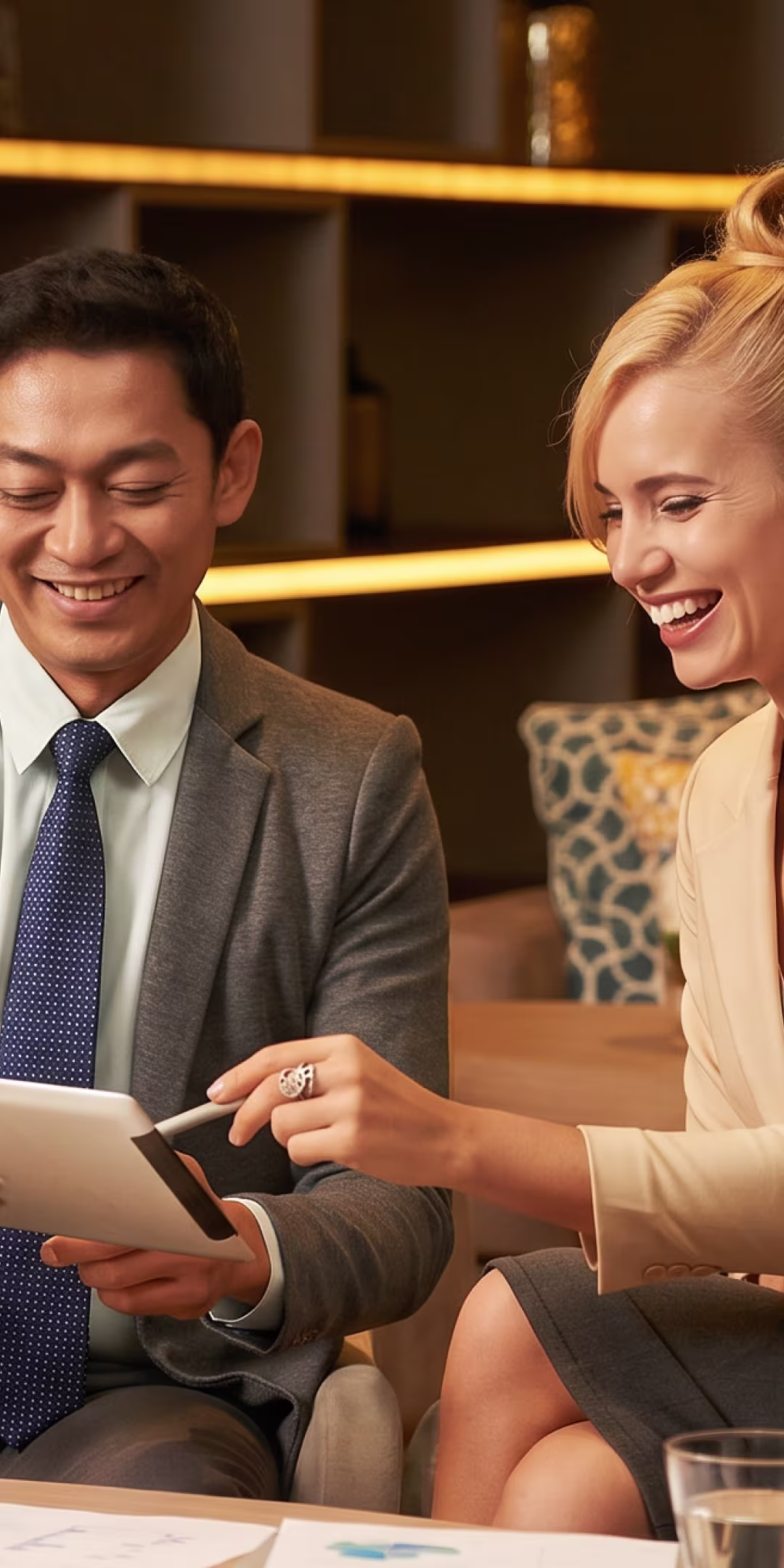 A man in a suit and tie and a woman in business attire smiling and discussing something on a tablet while sitting in a modern office lounge area.