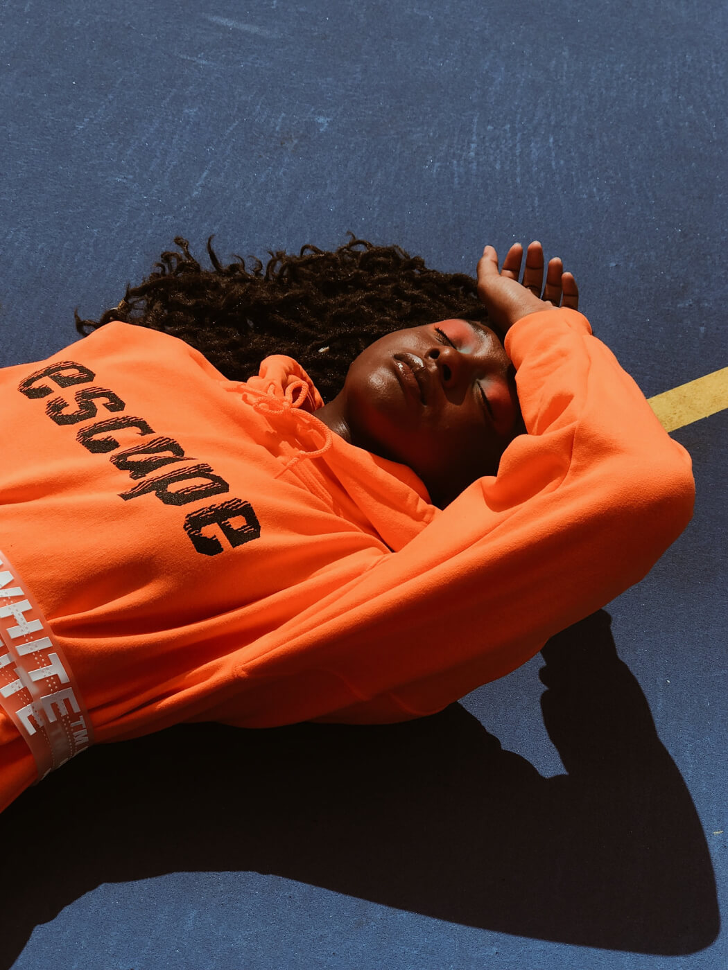 Person with dark skin and long curly hair lying on a blue surface with one arm resting over their forehead, wearing an orange hoodie with the word 'ESCAPE' in black letters and a white belt with text.