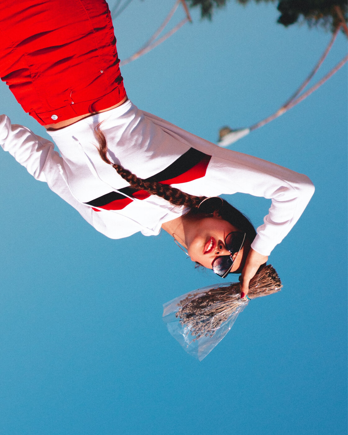Woman wearing a white long-sleeve shirt with red and black stripes and red pants, holding a wrapped bouquet of dried flowers against a clear blue sky, with her hair in a braid and wearing sunglasses.