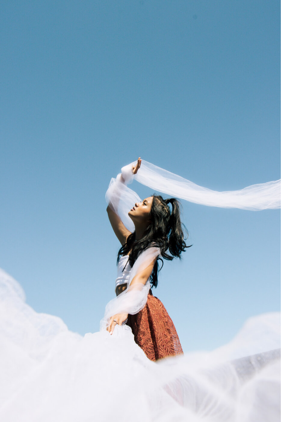 A woman with long dark hair tied in a ponytail stands against a clear blue sky, wearing a white top and a rust-colored lace skirt, holding and surrounded by flowing white translucent fabric that she lifts with one hand.