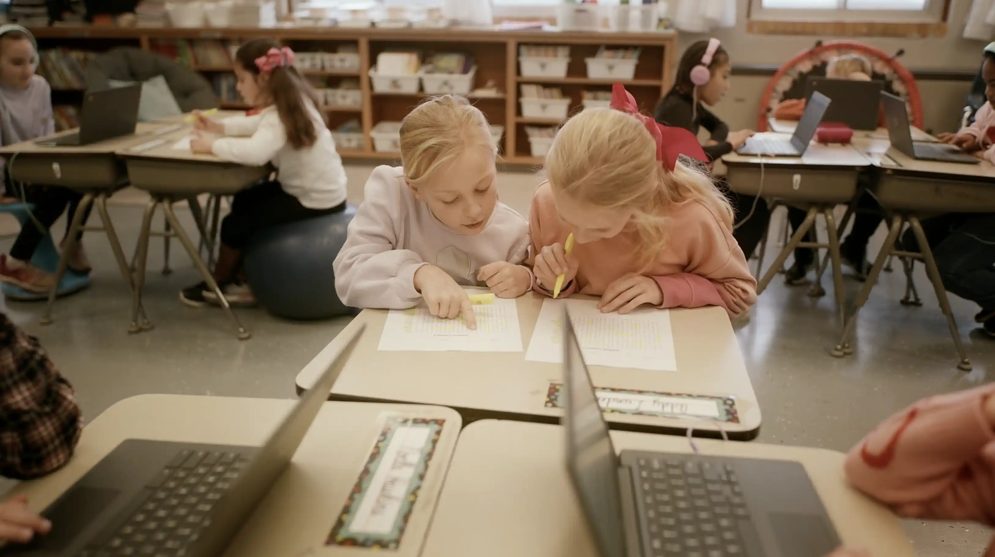 Two young girls sitting at desks in a classroom working together by highlighting text on printed papers, surrounded by other children using laptops and a bookshelf in the background.