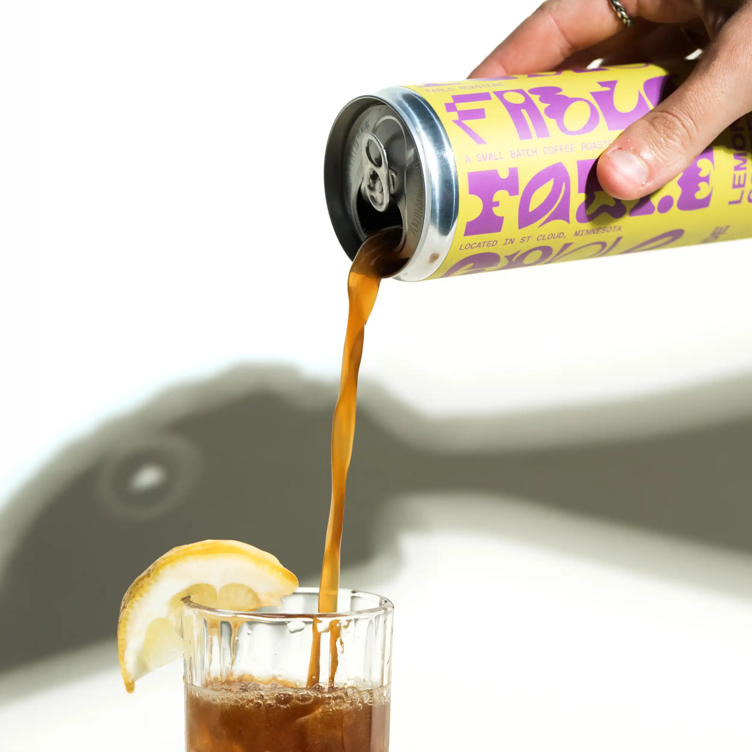 Hand pouring a dark beverage from a yellow and purple can into a glass with a lemon wedge on its rim and a fish-shaped shadow in the background.