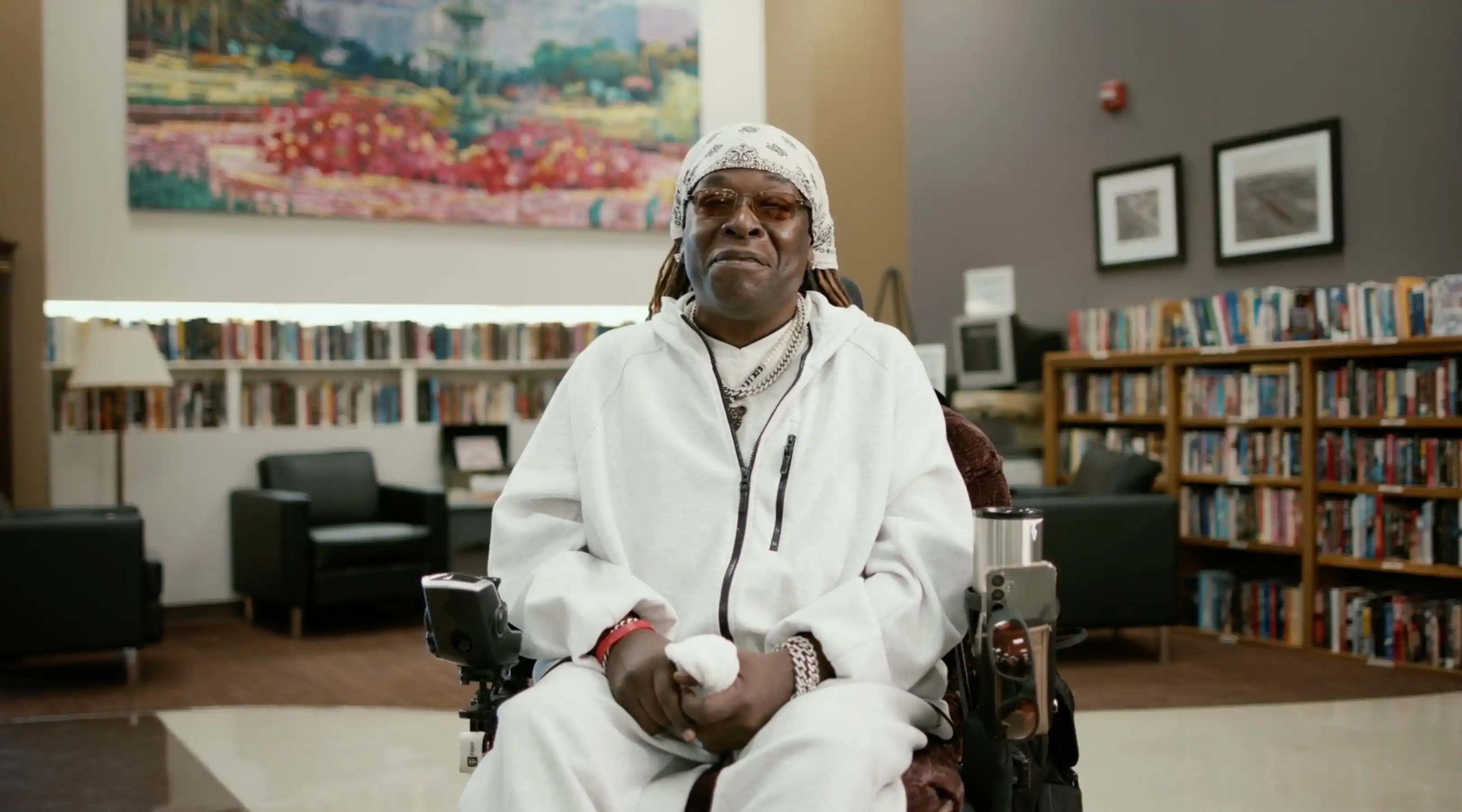A man wearing a white hoodie, white bandana, sunglasses, and chains sits in a motorized wheelchair inside a library with bookshelves and chairs in the background.