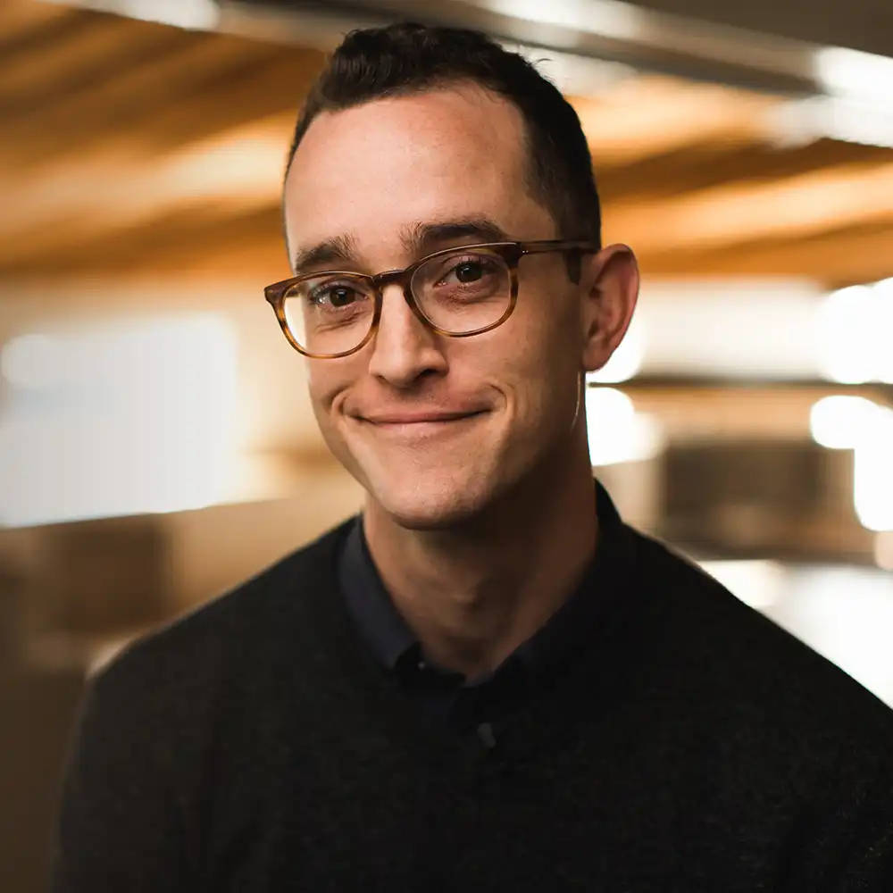 Smiling man with short dark hair and brown tortoiseshell glasses wearing a dark sweater in a warmly lit indoor setting with blurred background.