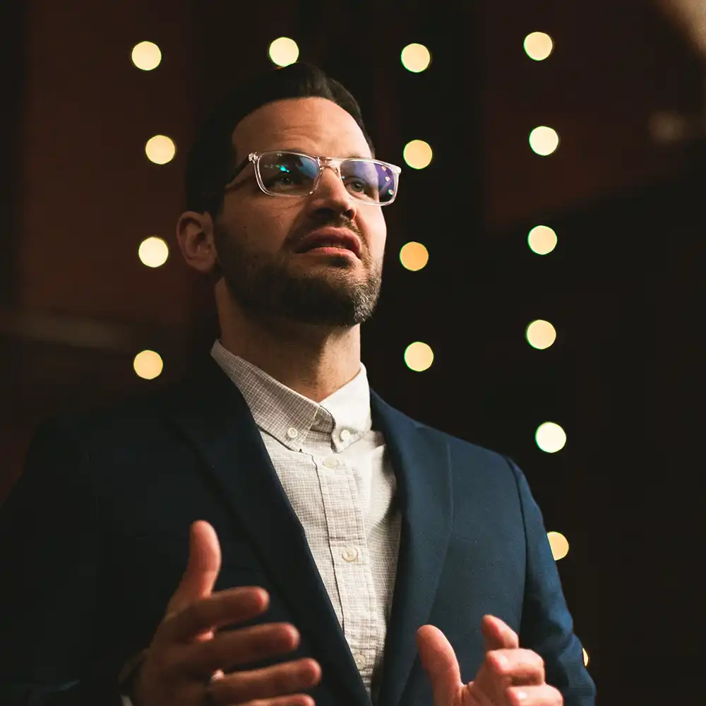 Man with beard and glasses speaking, wearing a dark jacket and light buttoned shirt, with blurred lights in the background.