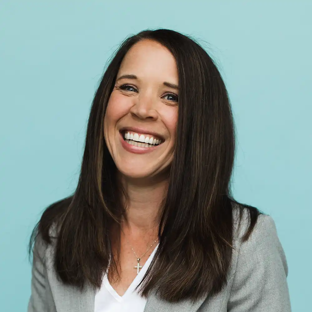 Smiling woman with long dark brown hair wearing a light grey blazer and a white top against a light blue background, with a silver cross necklace.