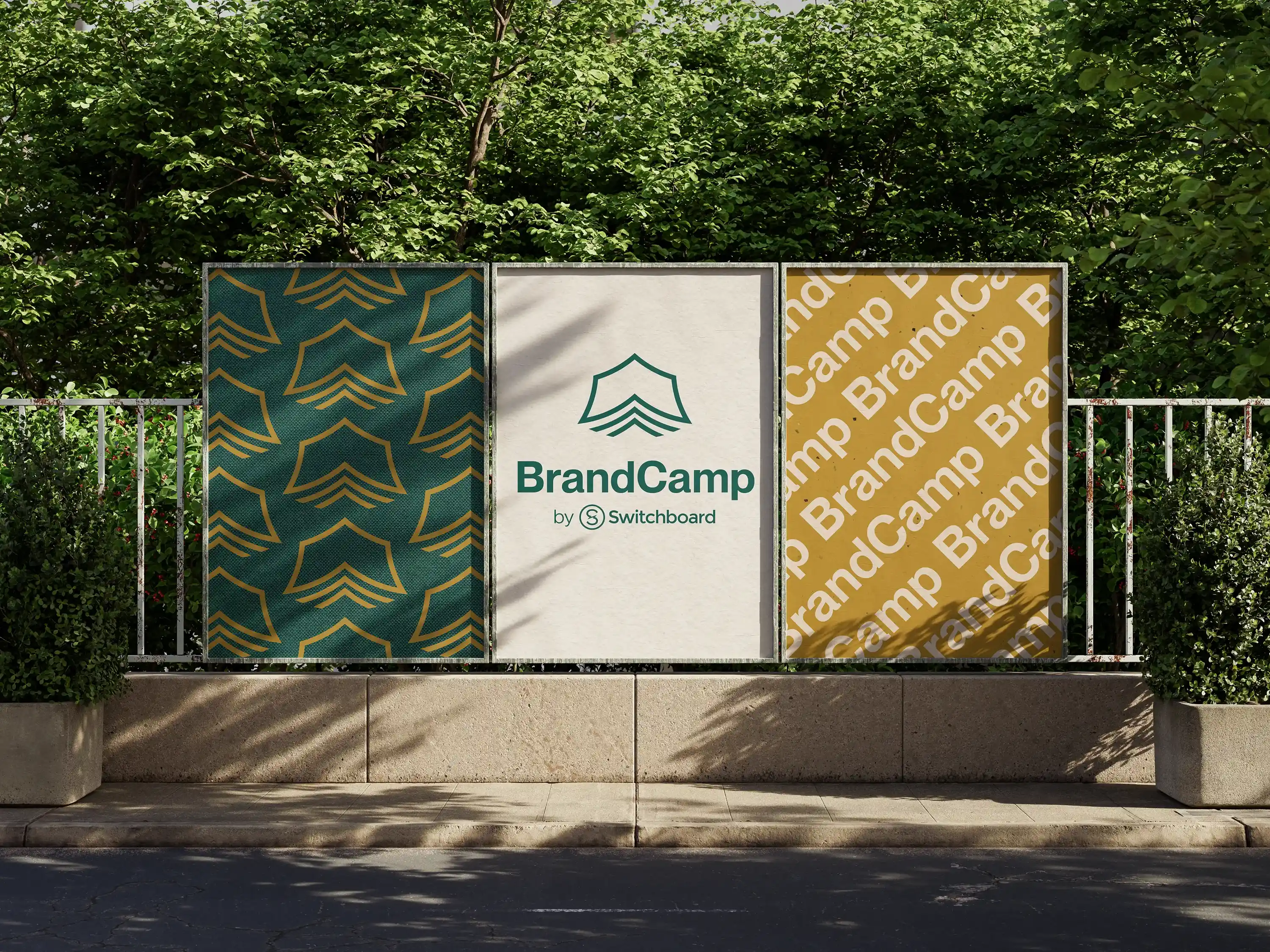 Three outdoor posters displayed side by side on a metal frame in front of greenery: left poster has a green background with a repeating gold shield-like pattern; center poster shows a green shield logo above the text "BrandCamp by Switchboard" on a white background; right poster has a gold background with repeated diagonal white text "BrandCamp."