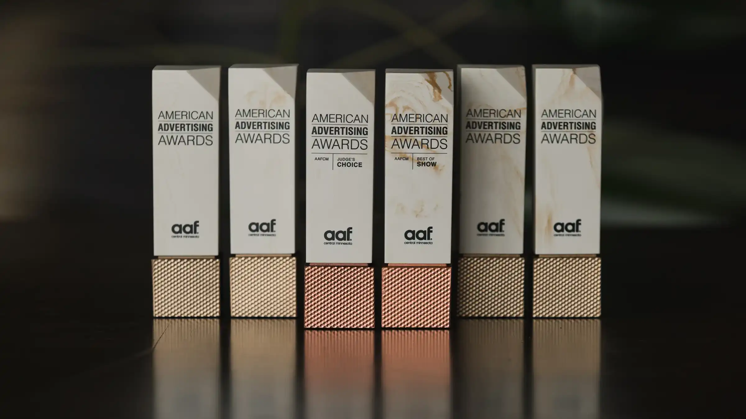Six American Advertising Awards trophies with rectangular tops and textured bases, two of which are labeled Judge's Choice and Best of Show by AAF Central Minnesota.