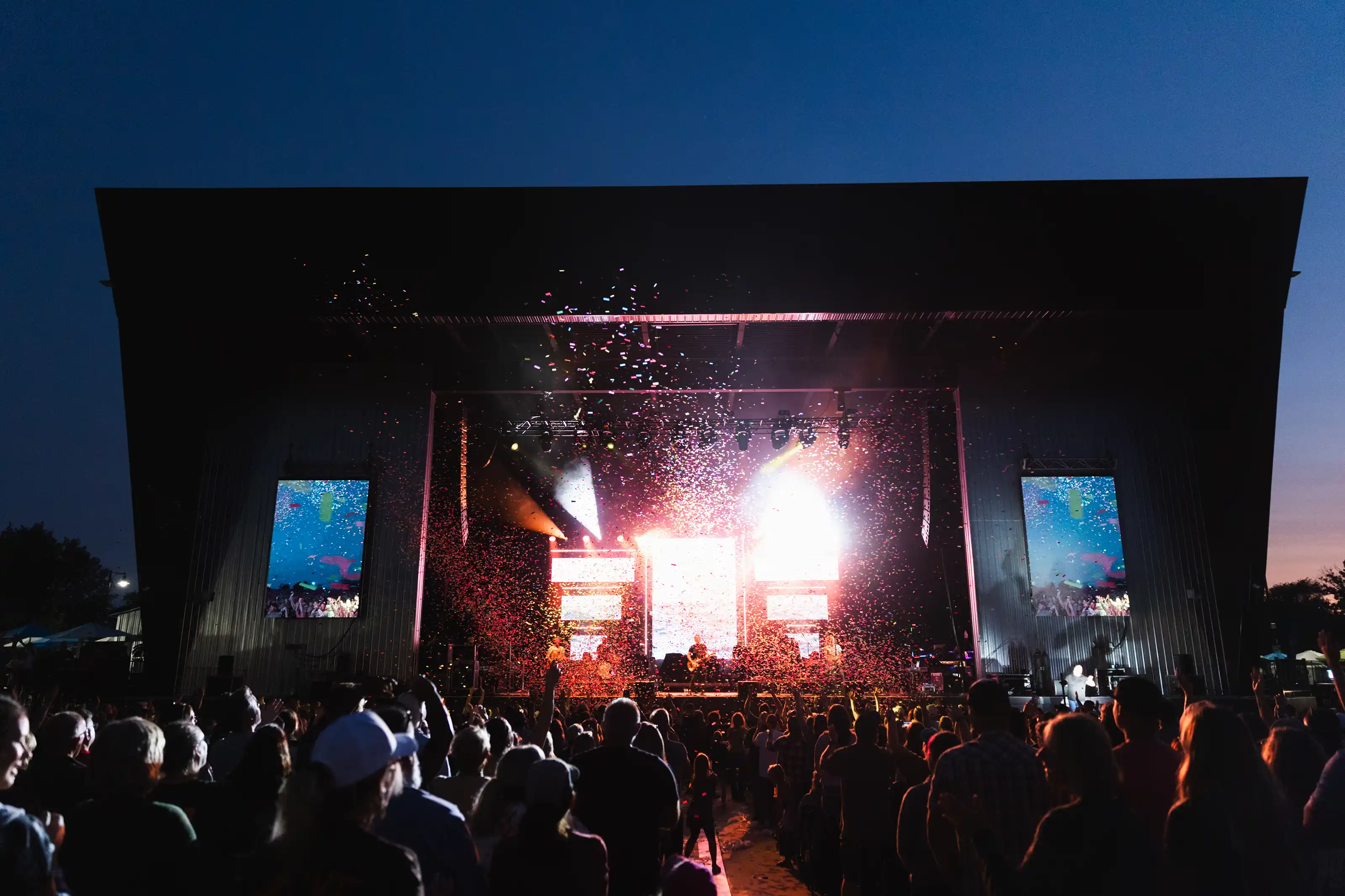 Crowd of people gathered at an outdoor concert stage at night with bright stage lights and confetti falling over the audience.