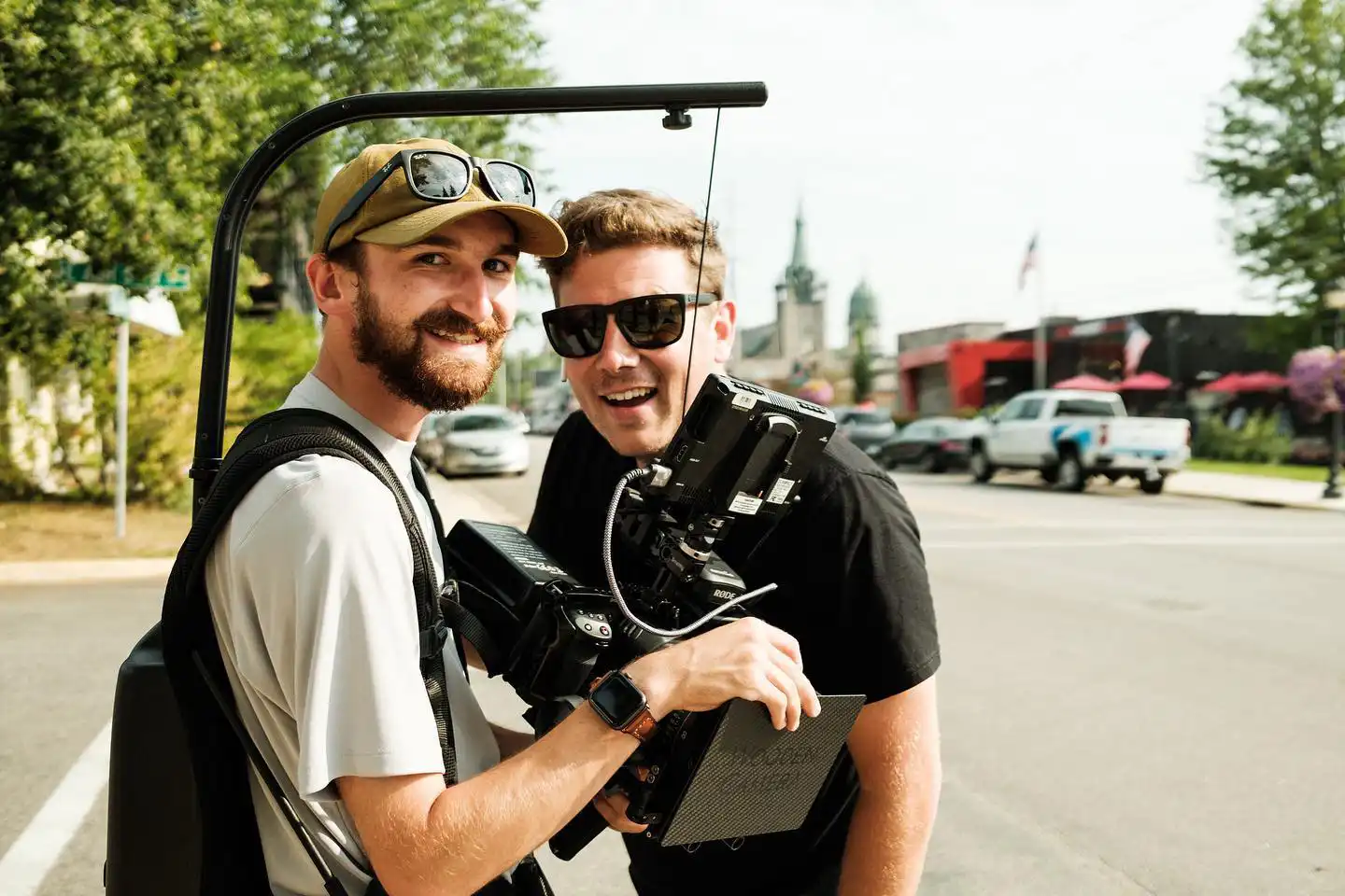 Two men outdoors, one wearing a tan cap and light shirt carrying professional video camera equipment with a stabilizer, both smiling at the camera on a sunny day with street and buildings in the background.