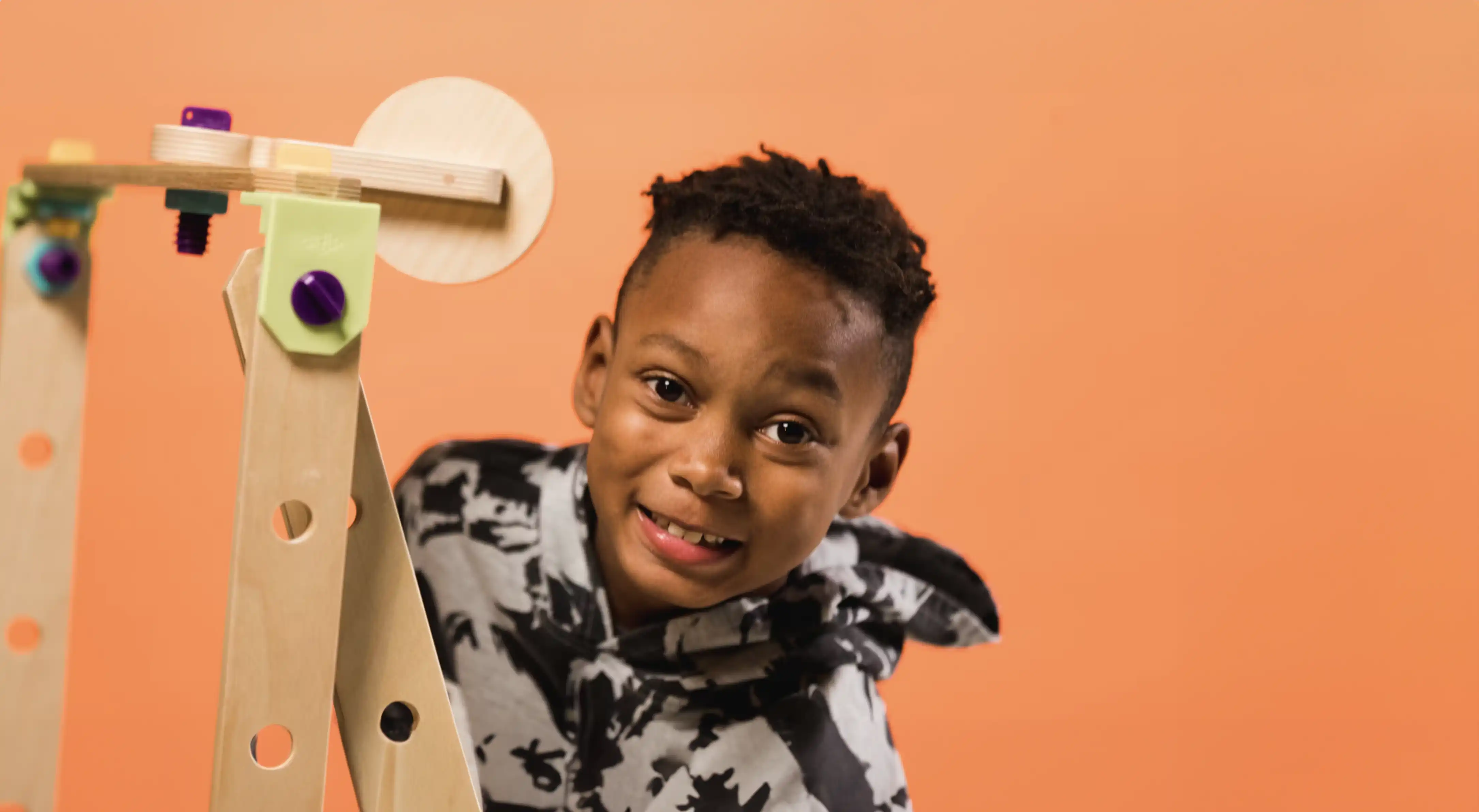 Boy with short curly hair smiling in front of an orange background, partially behind a wooden construction toy with purple and green connectors.