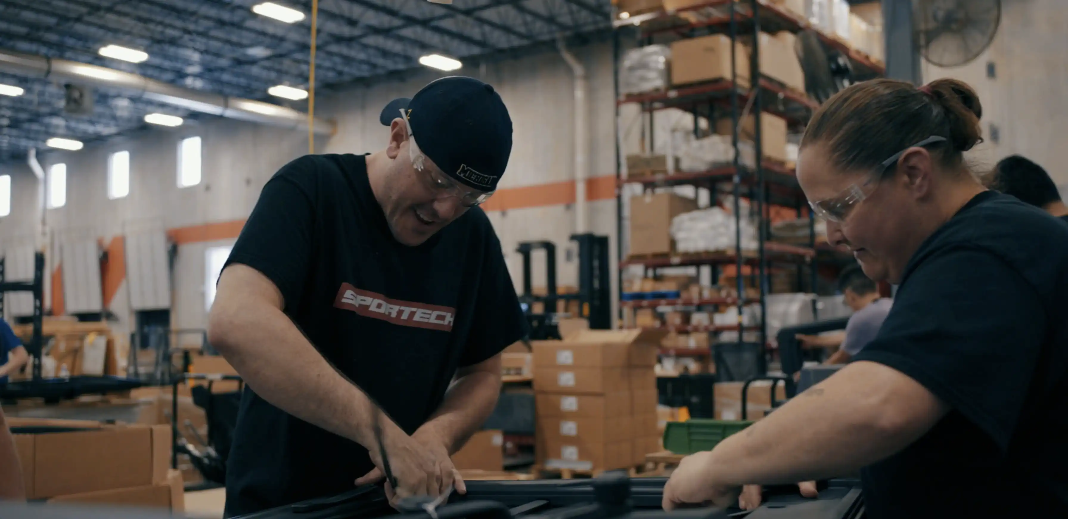 Two factory workers wearing safety glasses assembling or inspecting equipment inside a warehouse filled with shelves and stacked boxes.