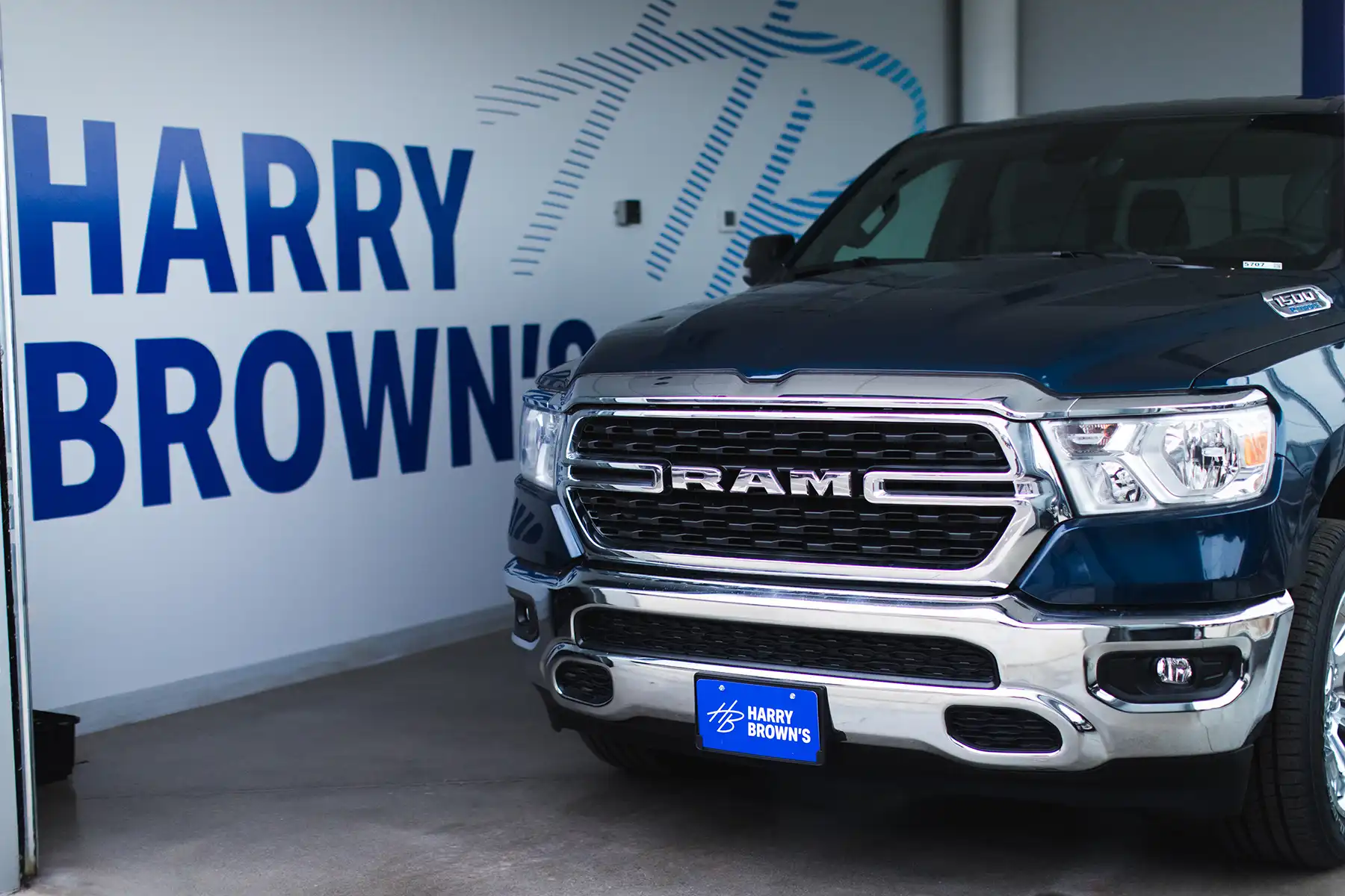 Front view of a dark blue RAM pickup truck with chrome grille and headlights parked indoors in front of a wall with large blue text 'HARRY BROWN'S' and a matching blue license plate with the same name and logo.