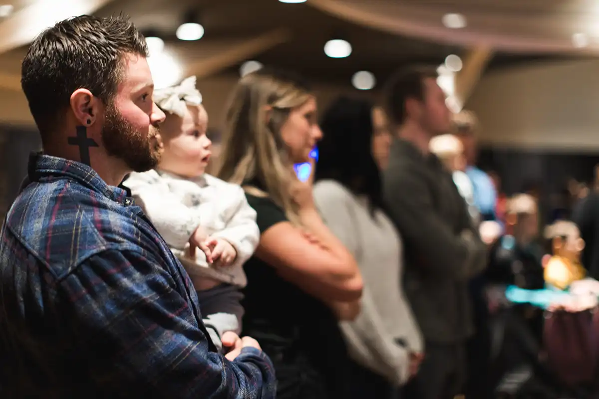 A man with a cross tattoo on his neck holds a baby wearing a bow headband while standing in a group of people indoors, all facing the same direction attentively.