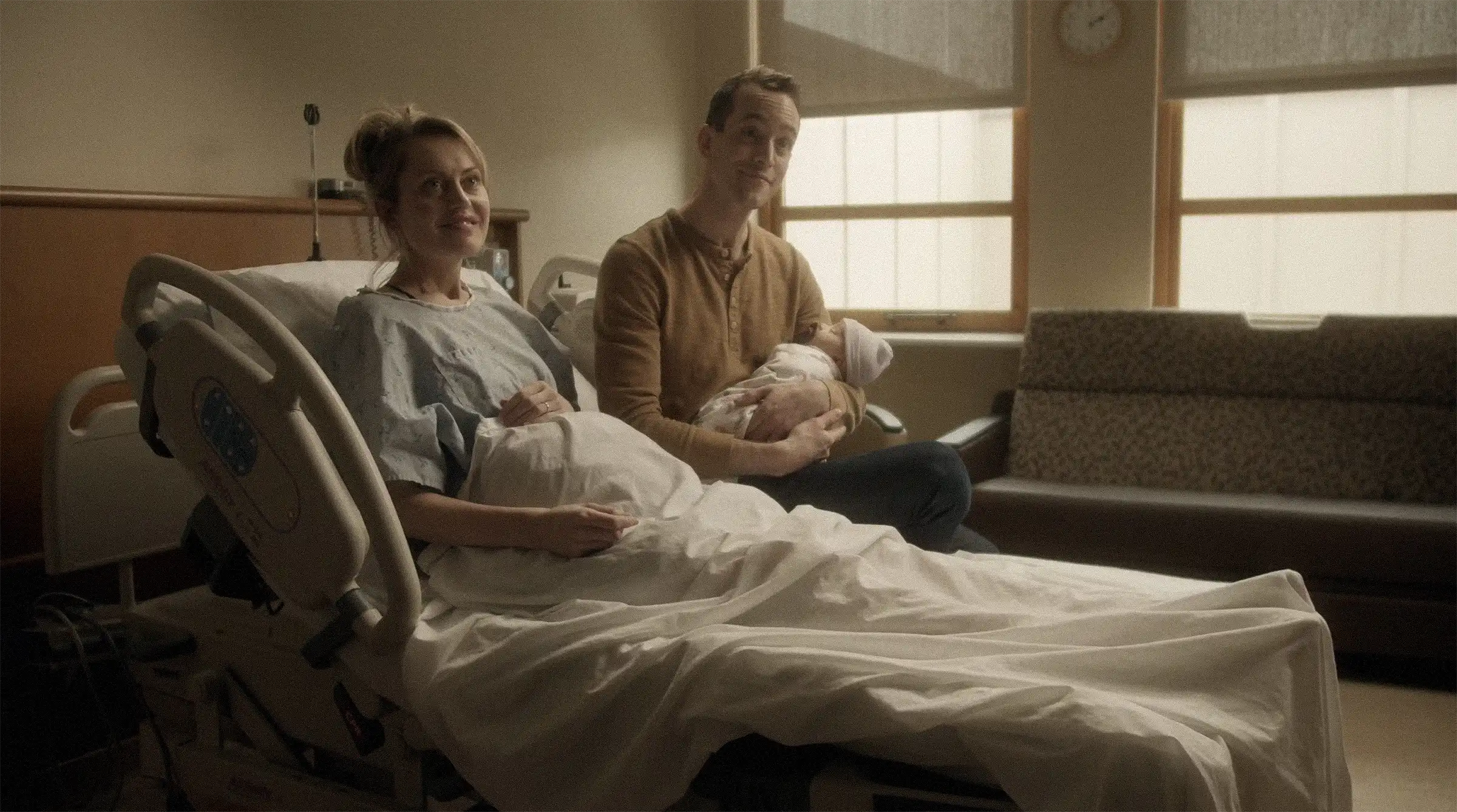 New mother lying in a hospital bed smiling, with a man sitting beside her holding a newborn baby wrapped in a blanket and wearing a hat.