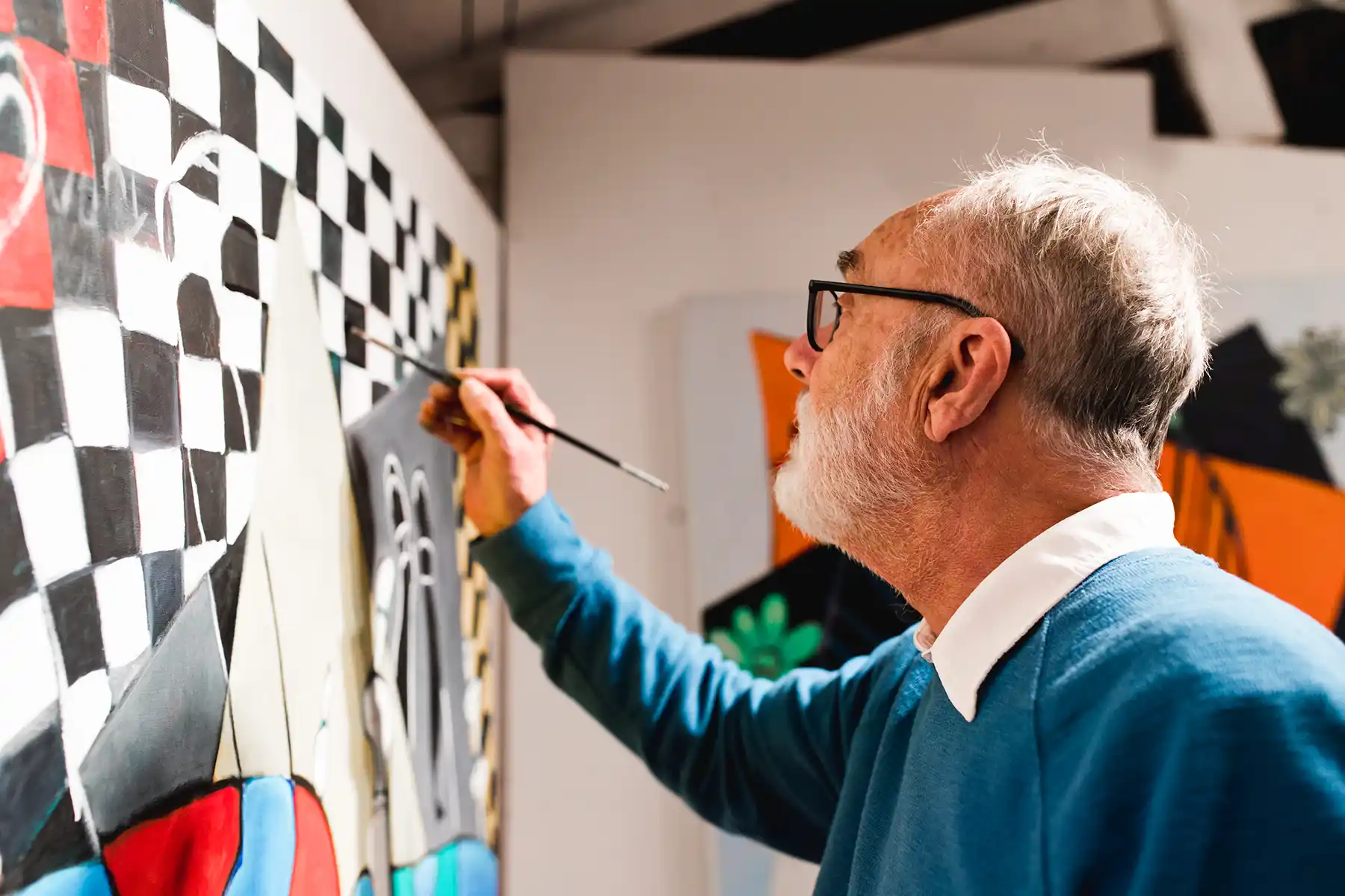 An elderly man with white hair and glasses painting a colorful abstract art piece featuring black and white checkered patterns and geometric shapes.