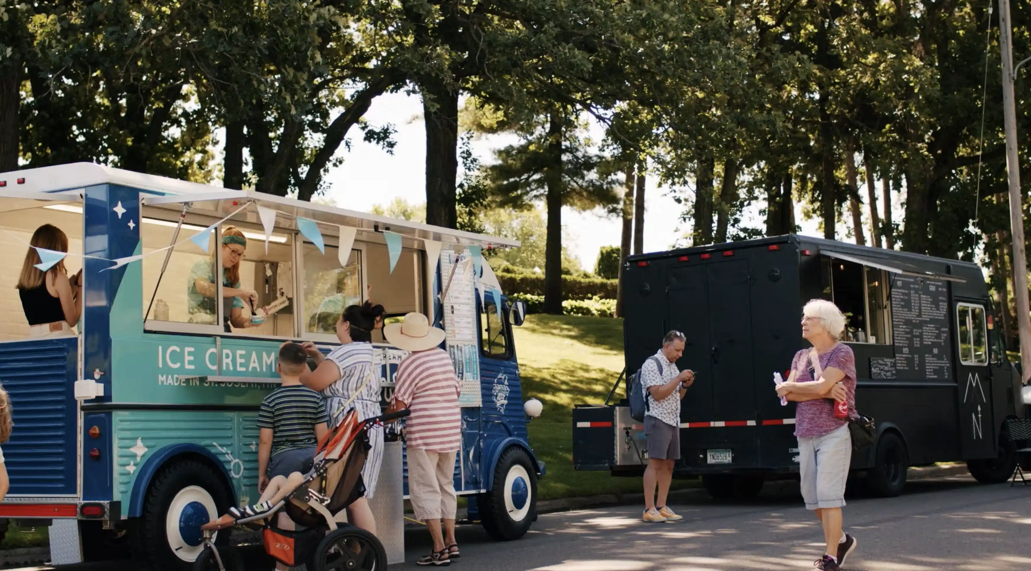 People gathered outdoors at two food trucks on a sunny day with trees in the background; one truck serves ice cream and has customers including a woman pushing a stroller, the other is black with a menu board, with a man standing nearby.