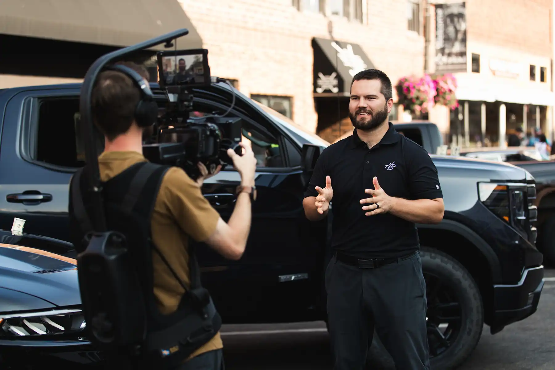 A man in a black polo shirt speaking in front of a black pickup truck while being recorded by a cameraman wearing headphones and a stabilization rig on a city street.