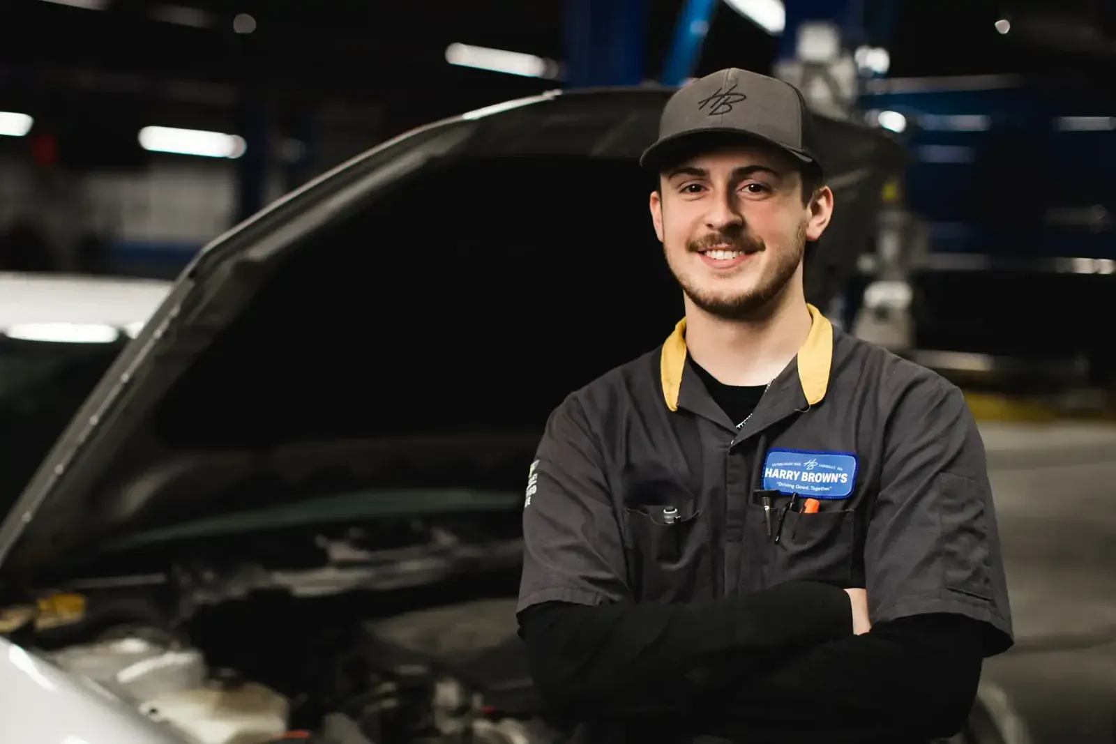 A male mechanic wearing a grey uniform with a Harry Brown's patch and a grey cap stands with arms crossed smiling in a garage with a car hood open behind him.