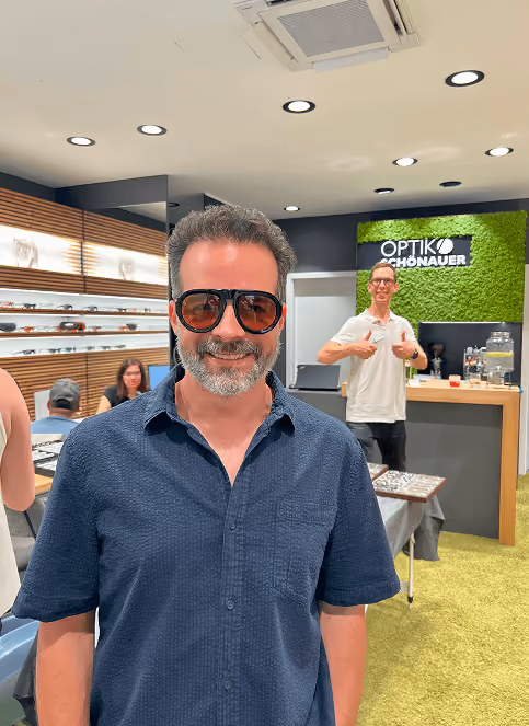 Mann mit Sonnenbrille in einem Optikergeschäft, lächelnd vor einem grünen Hintergrund, Verkäufer im Hintergrund.