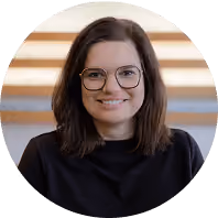 Lächelnde Frau mit dunklem Haar und Brille, trägt schwarzen Pullover vor gestreiftem Hintergrund.