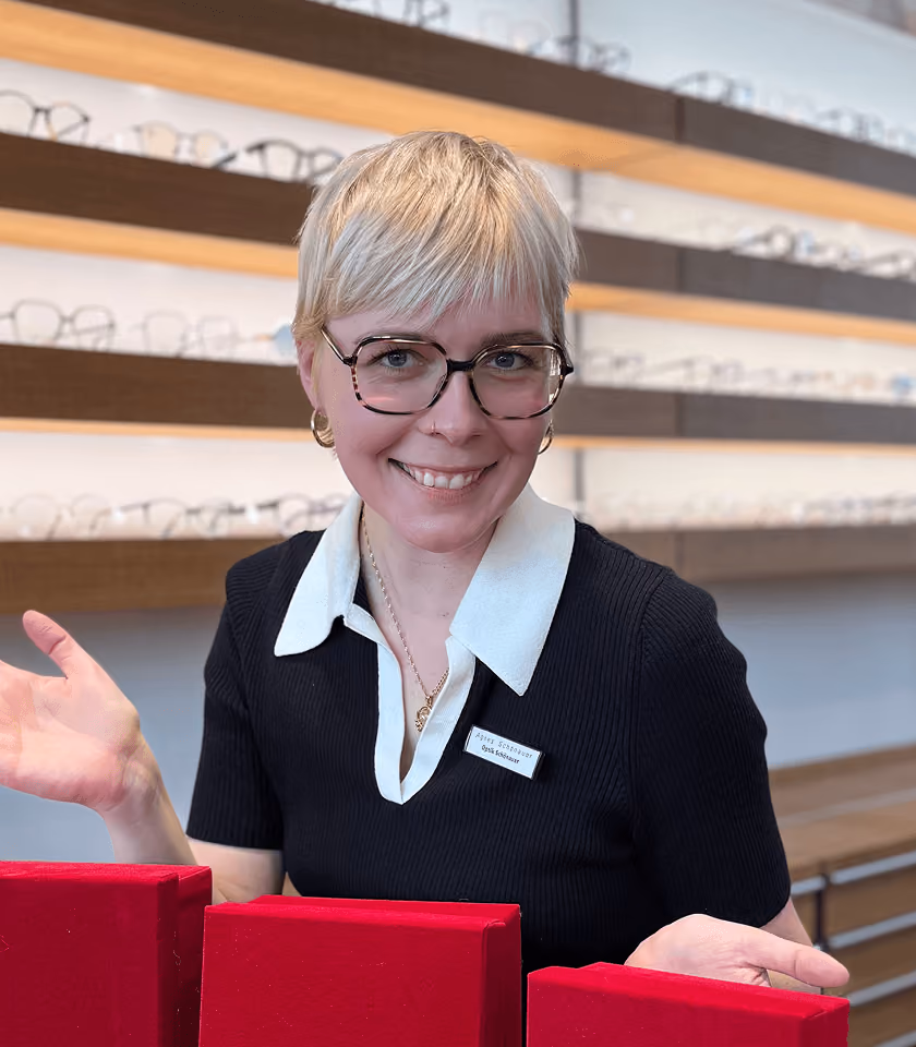 Blonde Frau mit Brille lächelt in einem Brillengeschäft, umgeben von Brillenregalen.