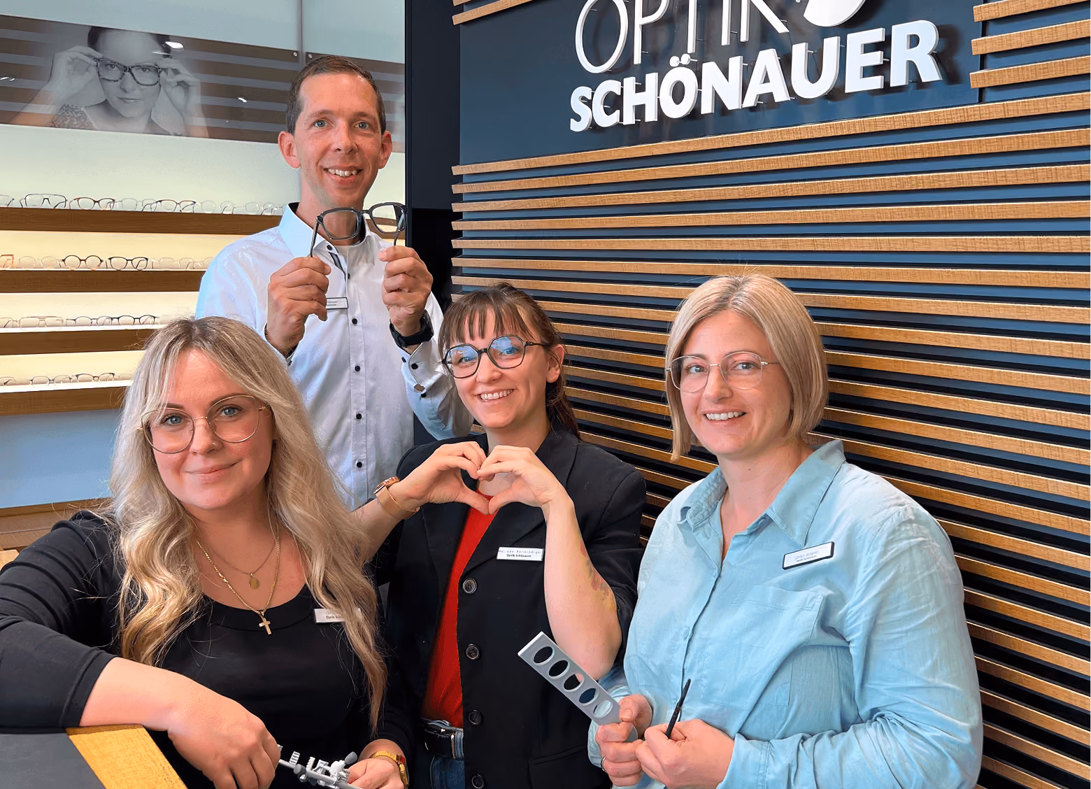 Vier Personen lächeln in einem Optikergeschäft, halten Brillen und ein Herzzeichen. Hintergrund mit Brillengestellen.