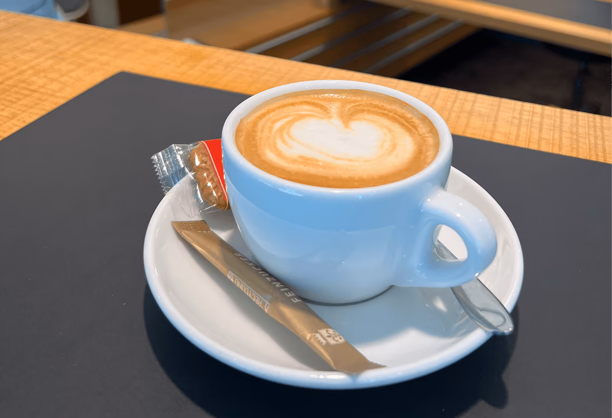 Cappuccino mit Herz-Latte-Art in weißer Tasse auf Untertasse, serviert mit Keks und Zuckertütchen.