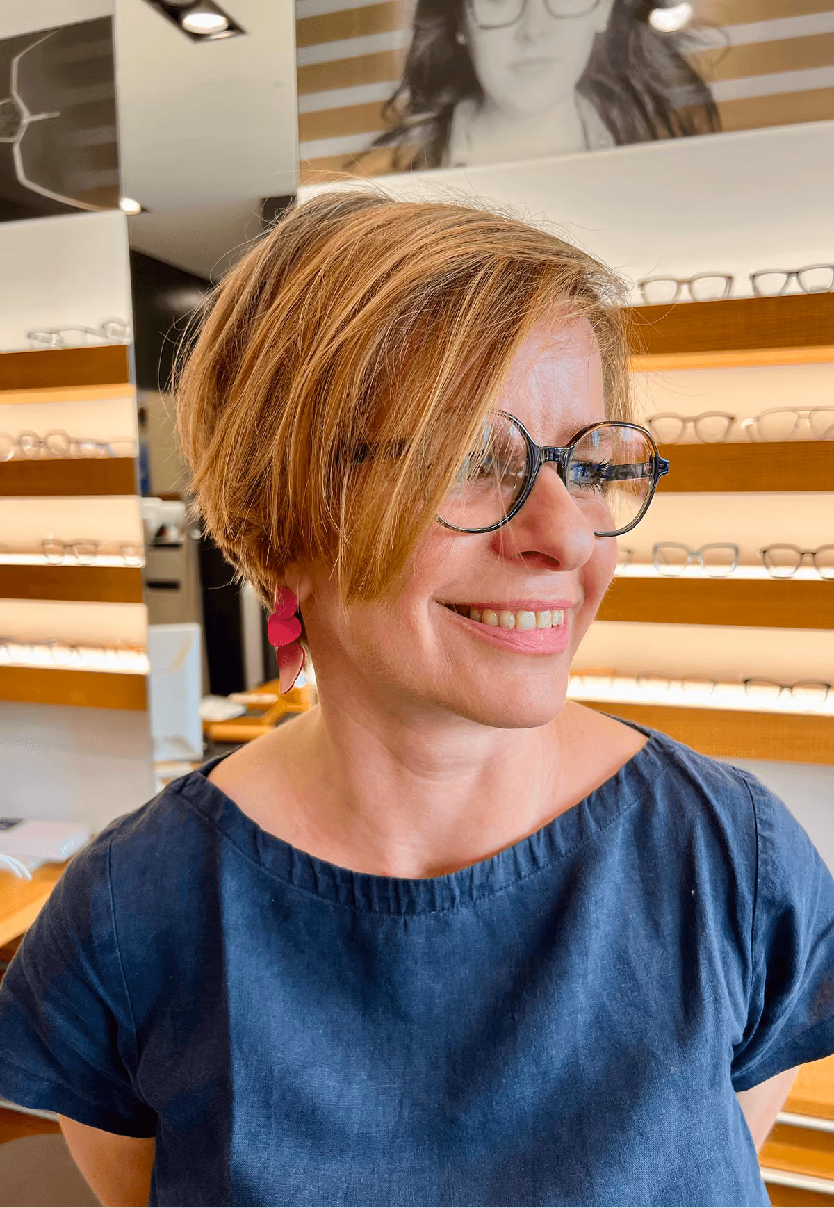 Frau mit kurzen blonden Haaren und Brille lächelt in Optikergeschäft.