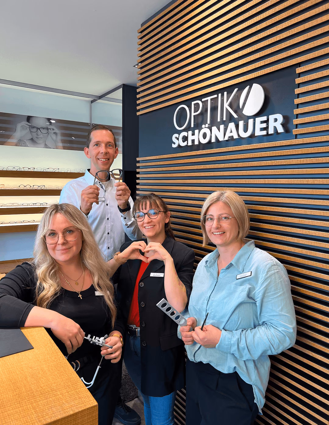 Vier Personen lächeln in einem Optikergeschäft, eine hält eine Brille, andere formen ein Herz mit den Händen. Hintergrund Holz-Dekor.