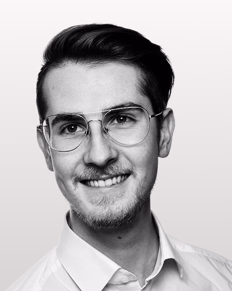 Porträt eines lächelnden jungen Mannes mit Brille, kurzem Haar und Hemd vor hellem Hintergrund.